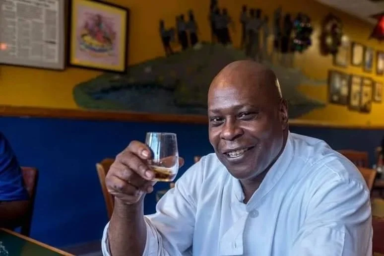 CHef Wayne Hooker A man smiling and holding a glass of whiskey in a restaurant with colorful wall decorations.