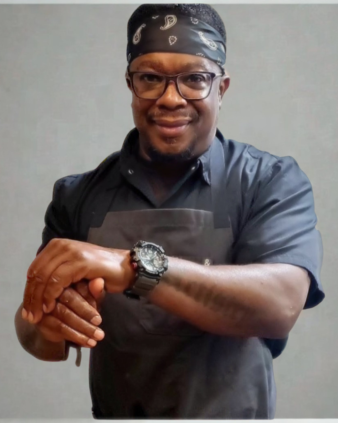A man with glasses and a bandana on his head, wearing a black shirt and apron, smiling and showing his watch, standing against a plain background. Chef Wayne G. Hooker Scholarship Board Member