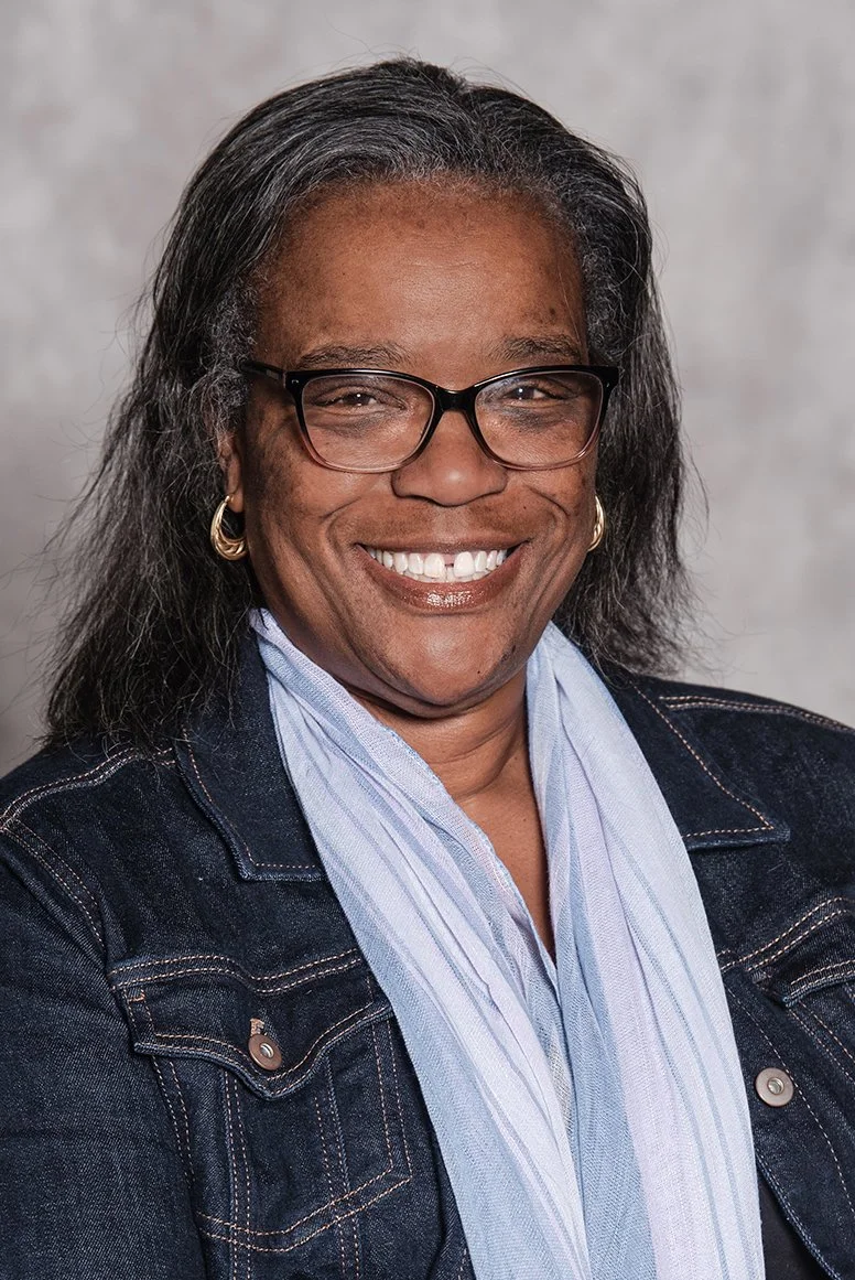 Chef Wayne G. Hooker Scholarship Board Member A smiling woman with glasses, earrings, and dark hair, wearing a denim jacket and light blue scarf against a neutral background.