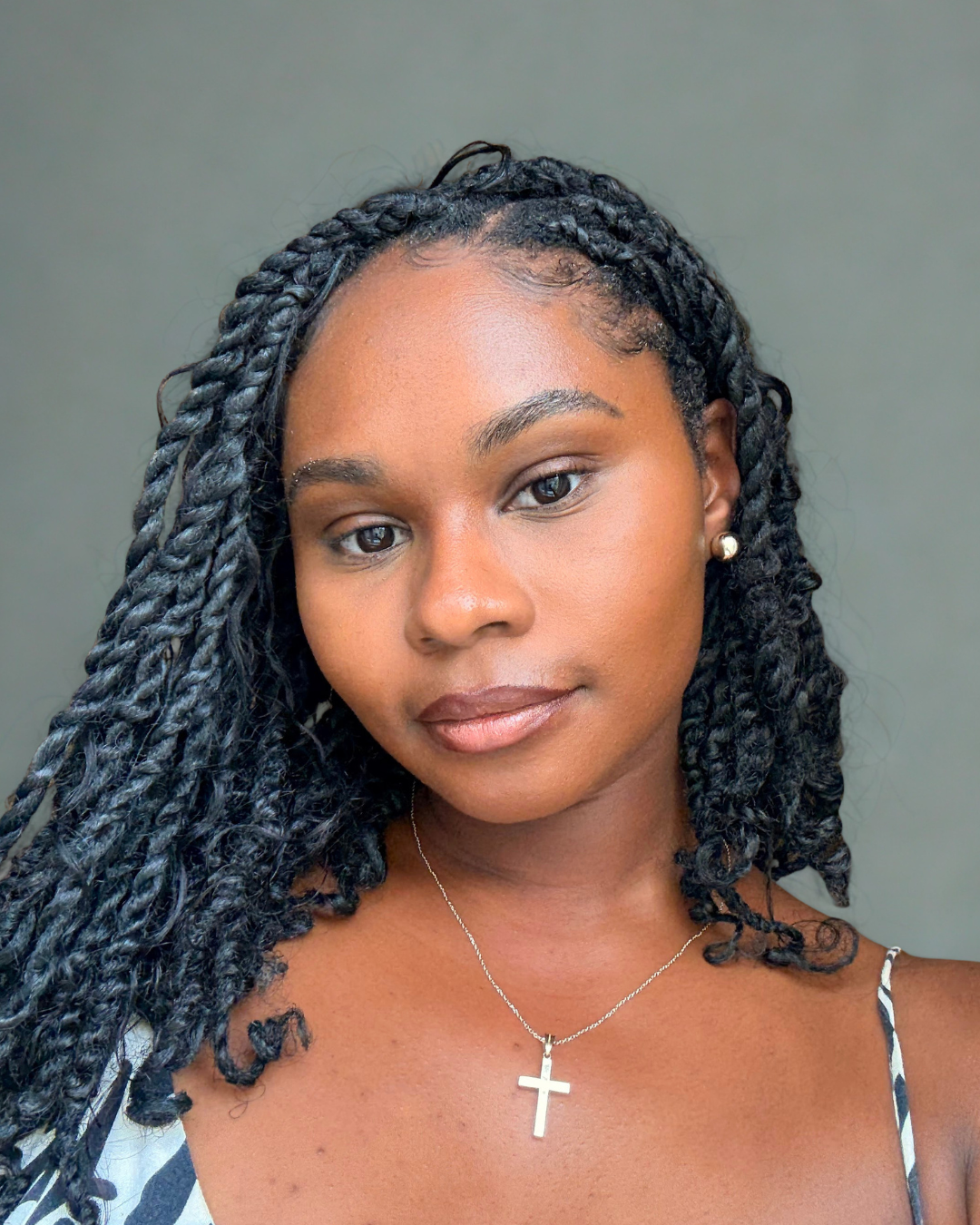 Chef Wayne G. Hooker Scholarship Board Member Close-up portrait of a woman with braided black hair, wearing pearl earrings, a cross necklace, and a sleeveless top with thin straps, standing against a plain gray background.