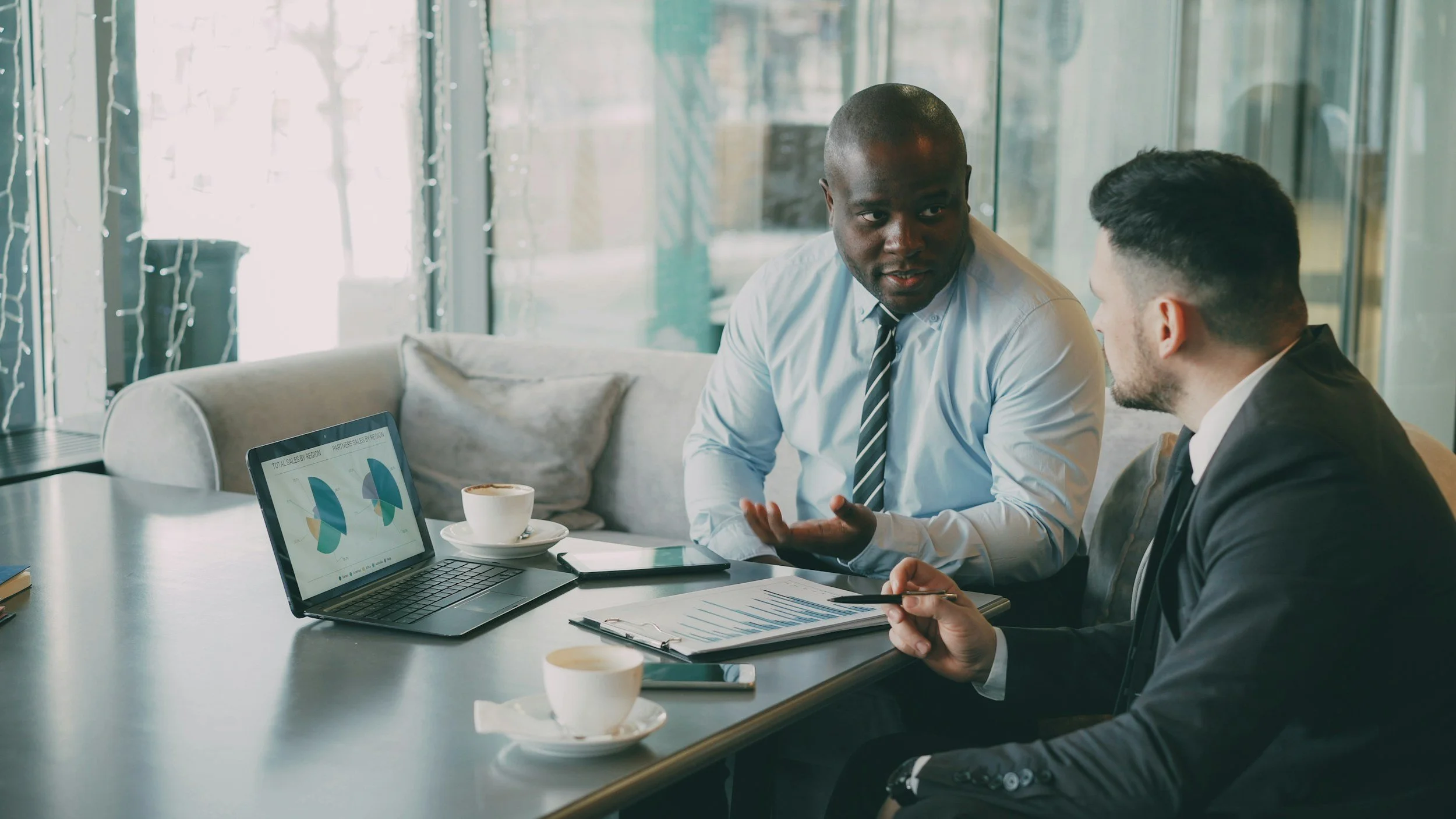 Two men in business attire having a discussion in a modern office with large windows, a laptop showing a chart, and coffee cups on the table.
