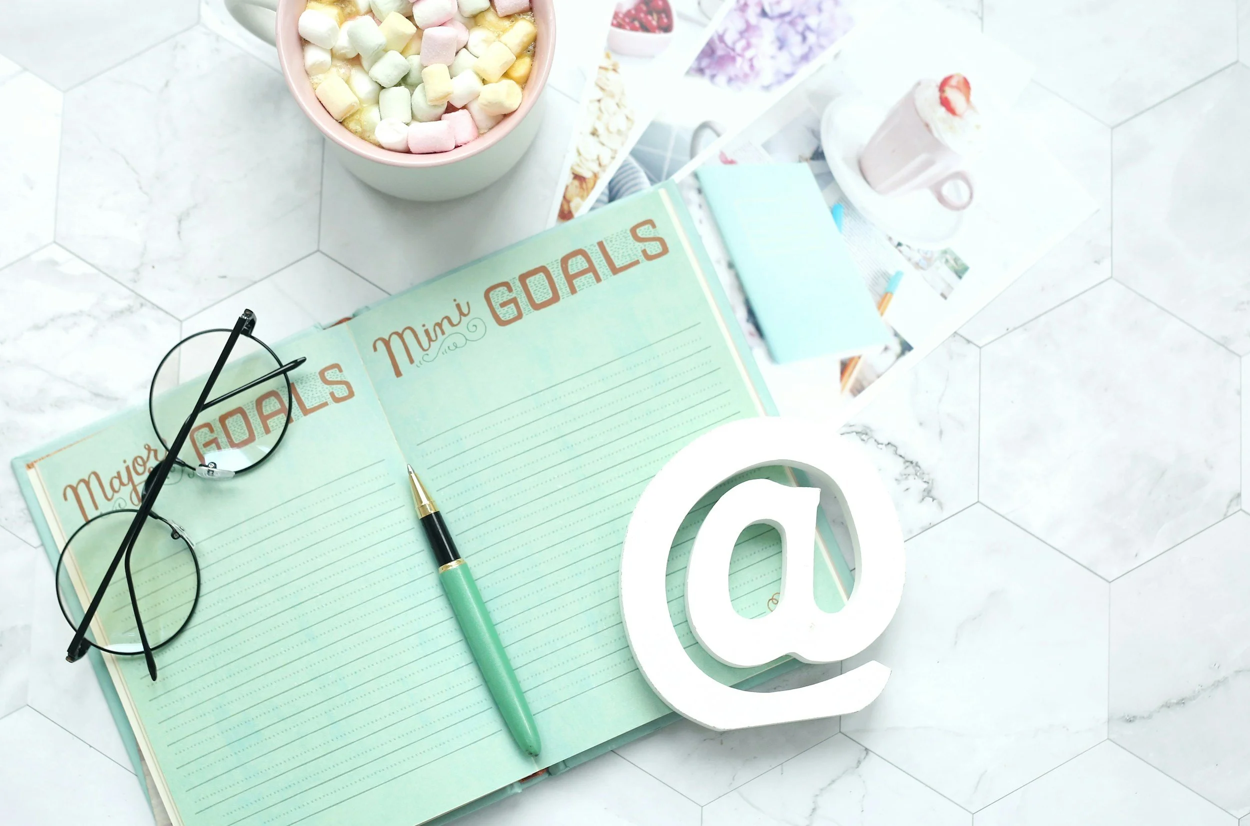 A desk scene with a light green notebook titled 'Major Mini Goals,' a black pair of glasses, a green pen, a white decorative letter 'O,' and a white mug with a pink beverage topped with whipped cream and a drizzle of red syrup. There is a pink bowl filled with colorful mini marshmallows on the desk and various photos and papers in the background on a white surface.