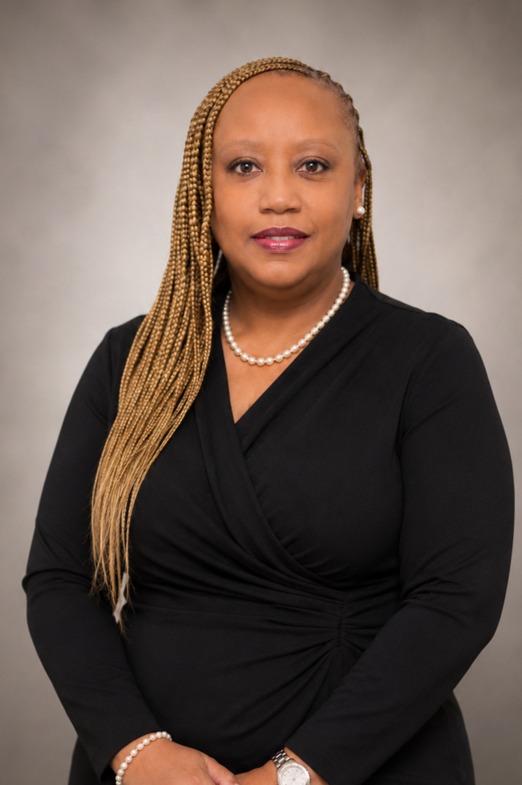 A woman with long blonde braids wearing a black dress, pearl necklace, pearl earrings, and a watch, posing against a neutral background.
