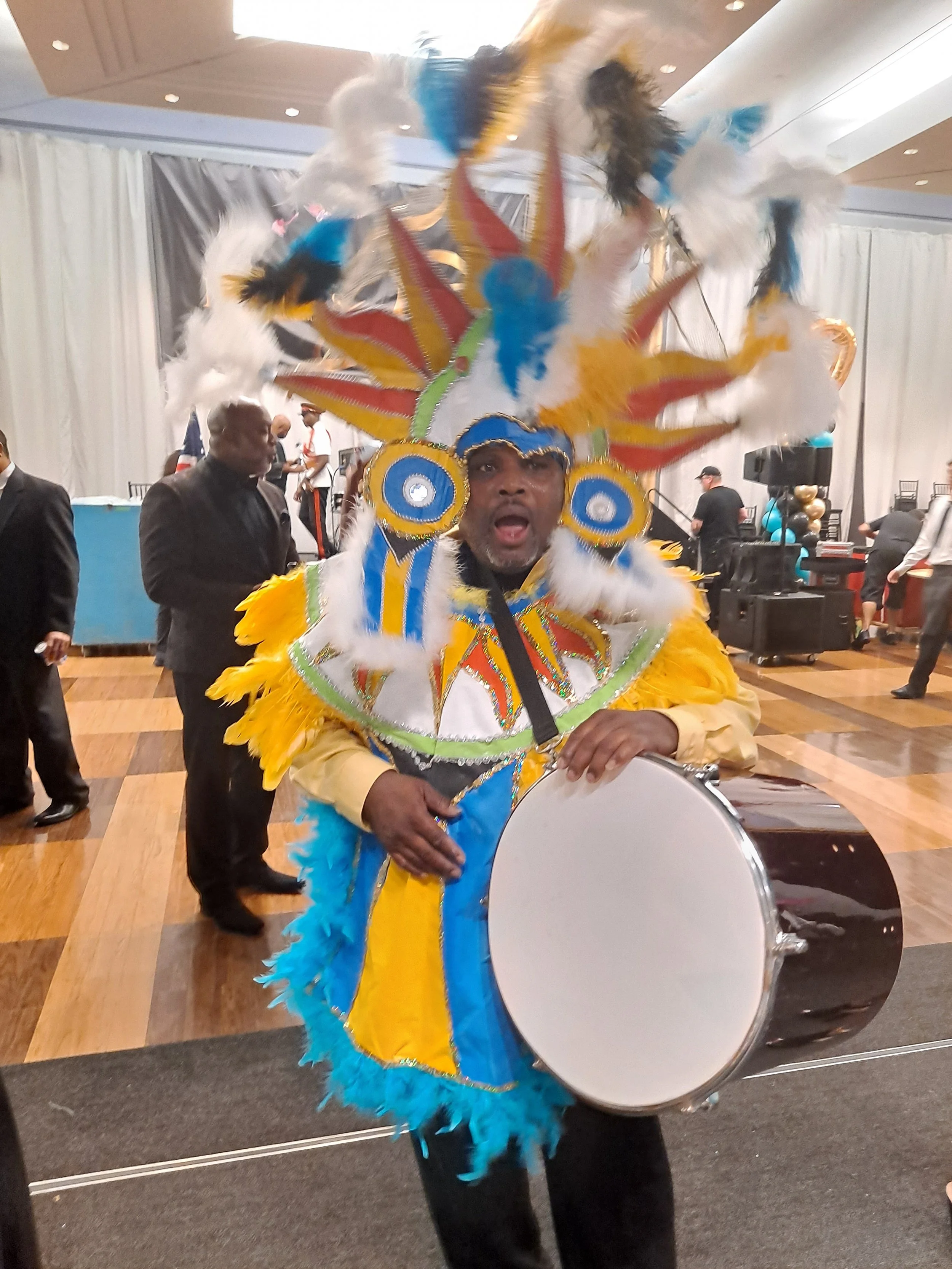 A person dressed in a colorful carnival costume with blue, yellow, white, and red feathers and embellishments, holding a large drum at an indoor event.