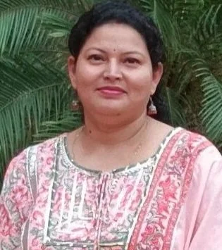 Woman with dark hair wearing a pink and white embroidered traditional outfit, standing outdoors with green foliage in the background.