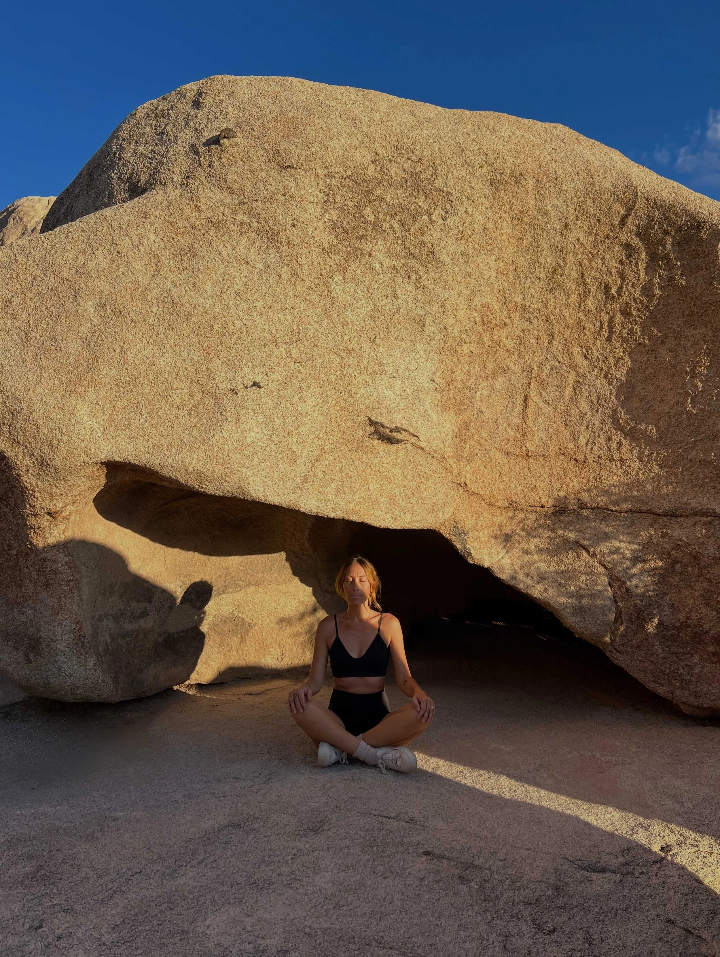 Une femme en position de méditation assise dans une pose de yoga devant une formation rocheuse en plein air, sous un ciel bleu.