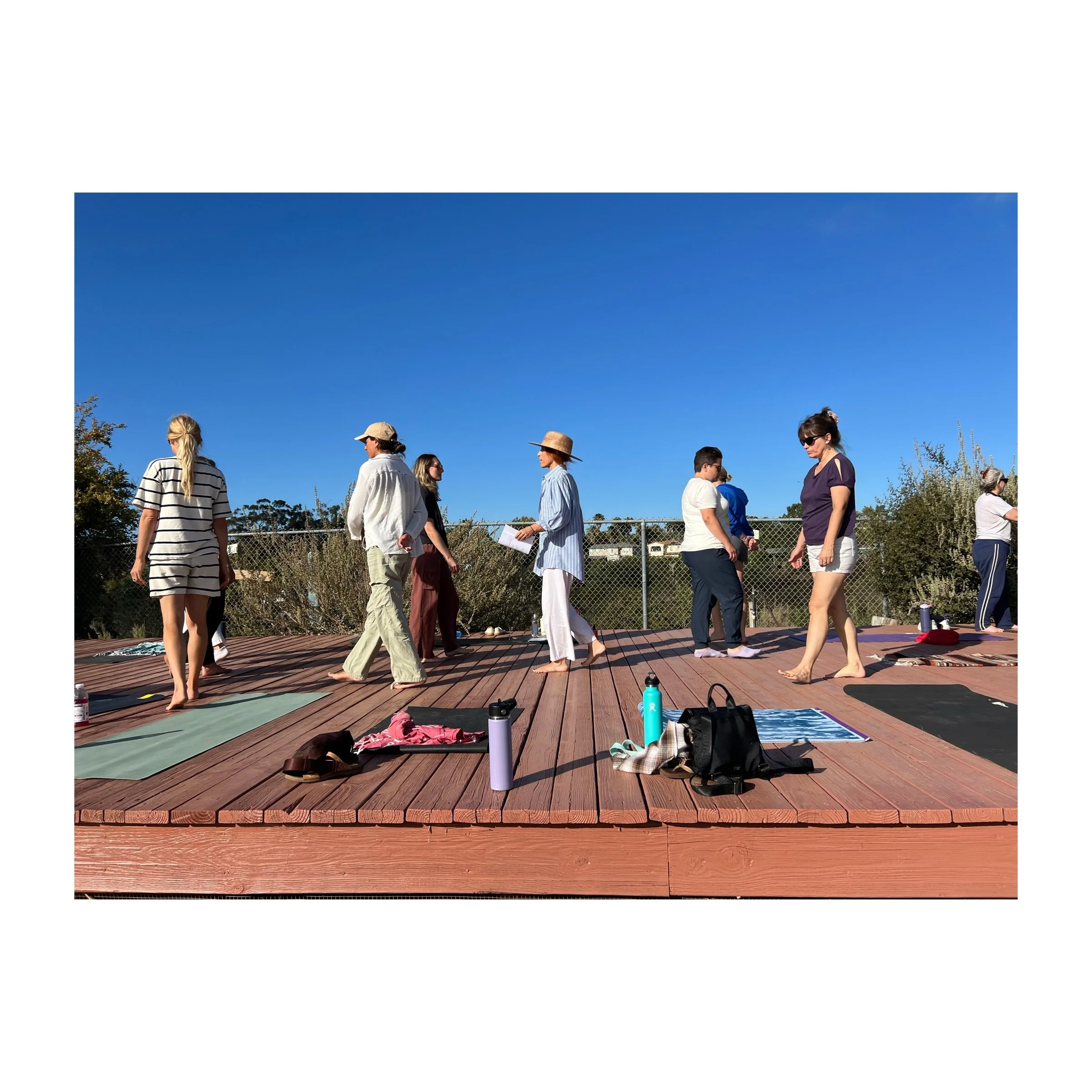Groupe de personnes faisant du yoga ou de la méditation sur une plateforme en bois en plein air, sous un ciel bleu clair, avec des sacs et des bouteilles d'eau à proximité.