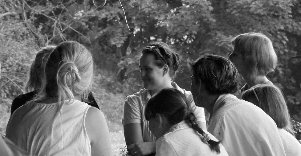 Groupe de personnes, principalement des femmes, en discussion en plein air avec une végétation en arrière-plan.
