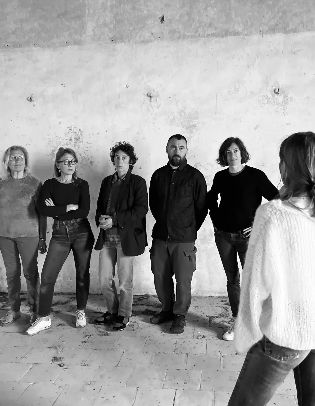 Groupe de six personnes debout devant un mur en pierre dans une pièce nue, en train d'écouter ou de regarder une femme à droite qui parle ou présente quelque chose.