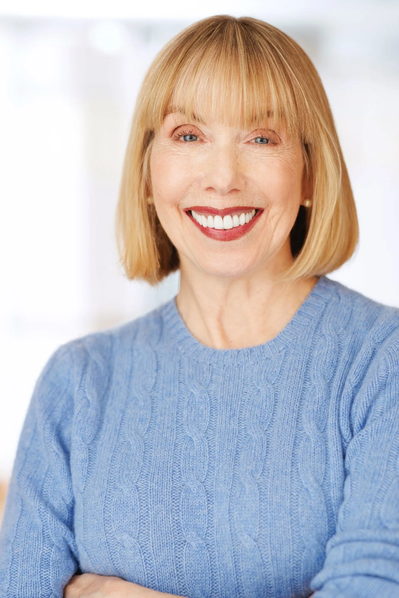 Close-up of smiling middle-aged woman with straight, shoulder-length blond hair and blue eyes, wearing a light blue sweater, with arms crossed indoors.