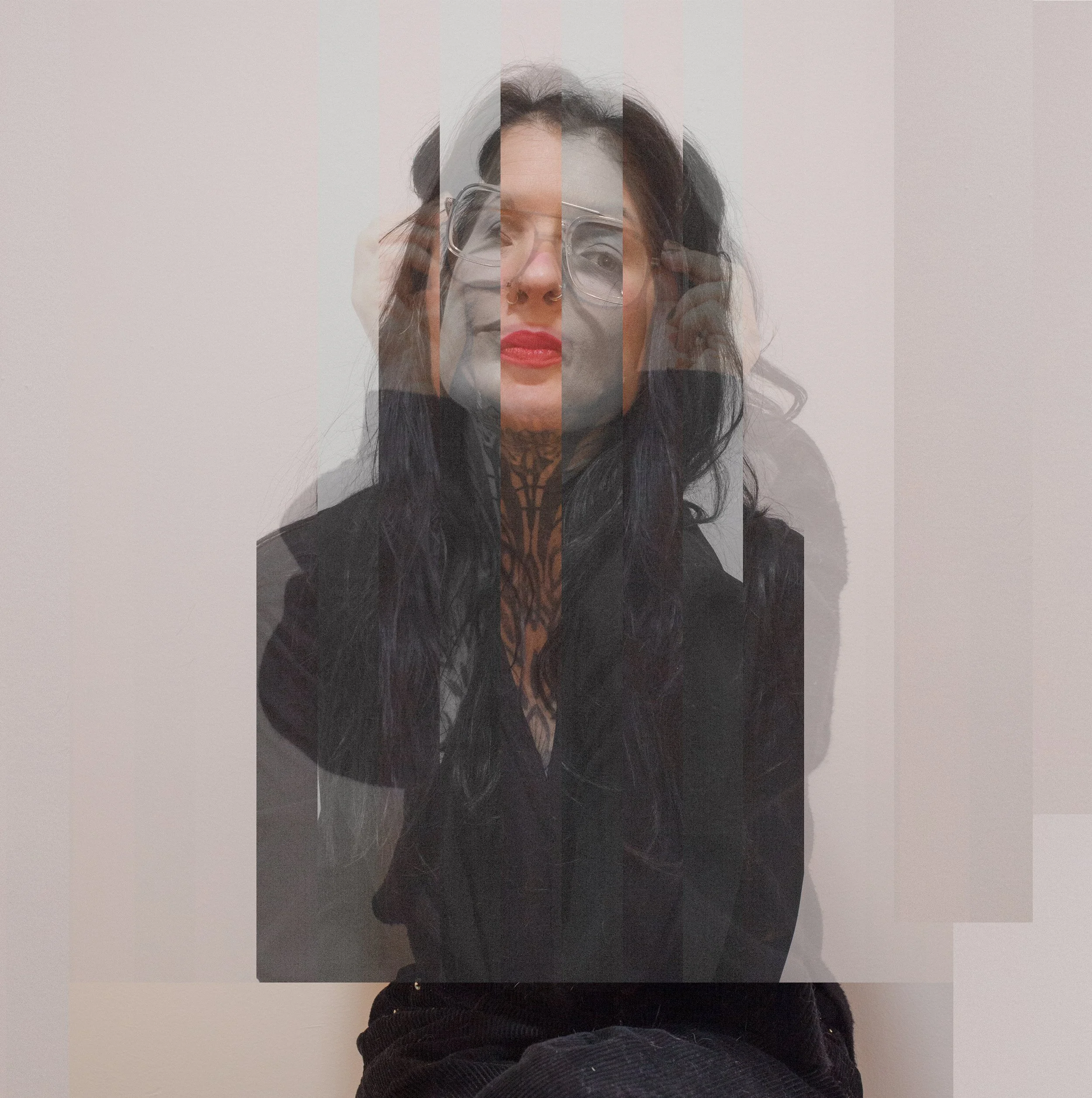 Tiffany Kelley - A woman with long dark hair, glasses, red lipstick, and a nose piercing, wearing a black top with lace detail, is sitting against a plain white or light-colored wall.