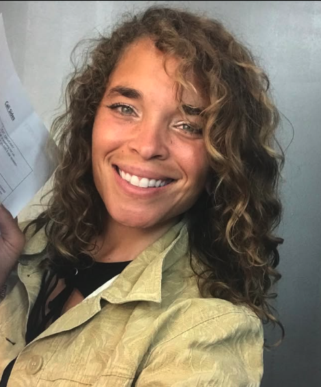 MAYA ROCK - A woman with curly blonde hair and blue eyes smiling in front of a gray wall, holding a paper in her left hand.