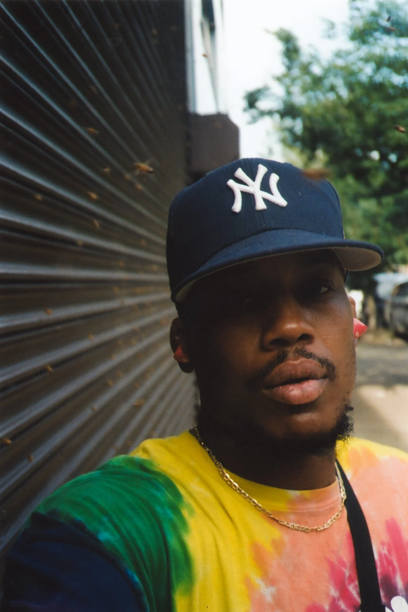 MUTAURWA MAPONDERA  - A man with a beard and dark skin wearing a black New York Yankees baseball cap and a multicolored tie-dye shirt, standing outdoors near a metal fence with green trees in the background.