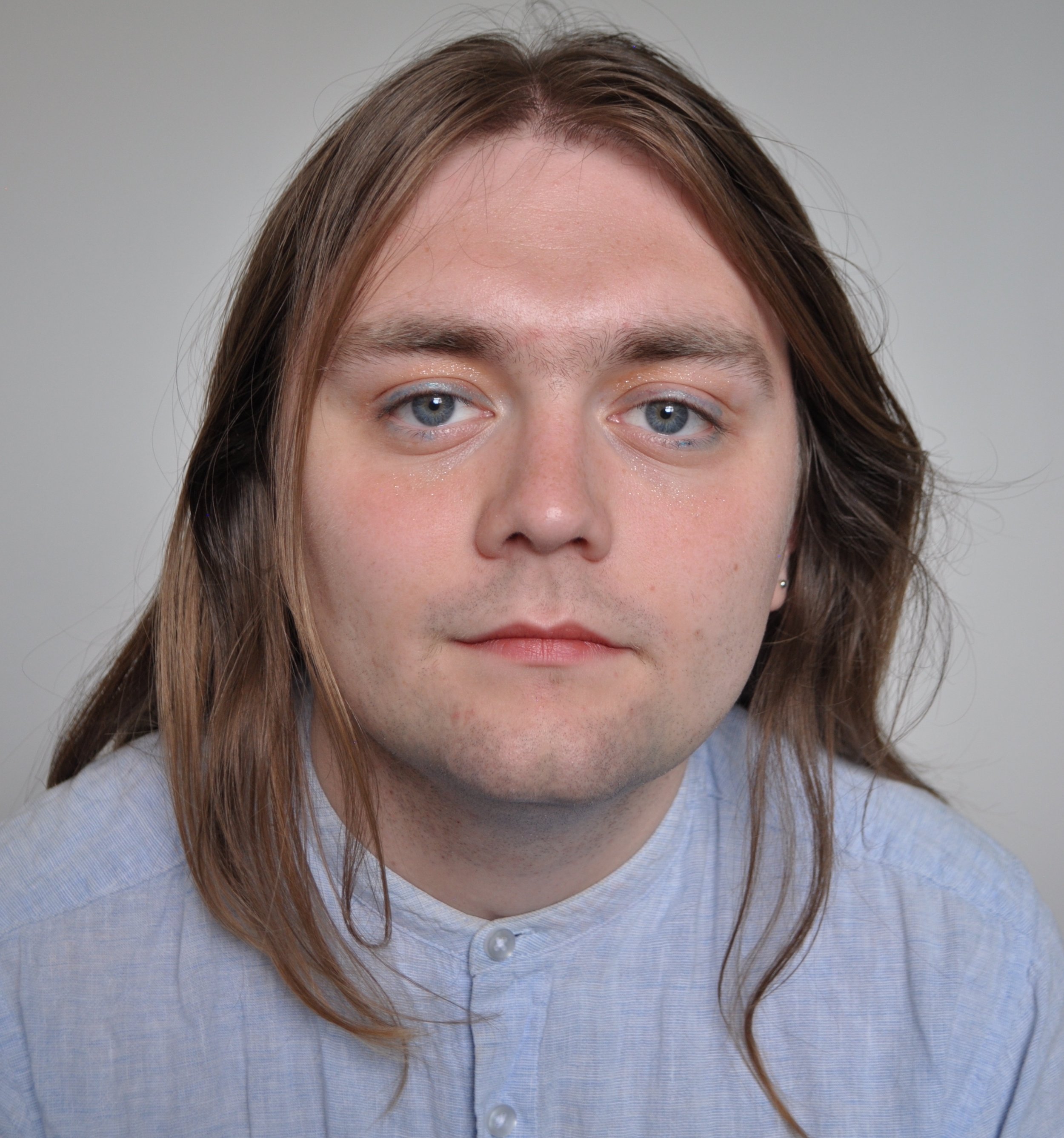 Jack Warren - Close-up of a person with long brown hair, blue eyes, and fair skin, wearing a light blue collared shirt against a neutral background.