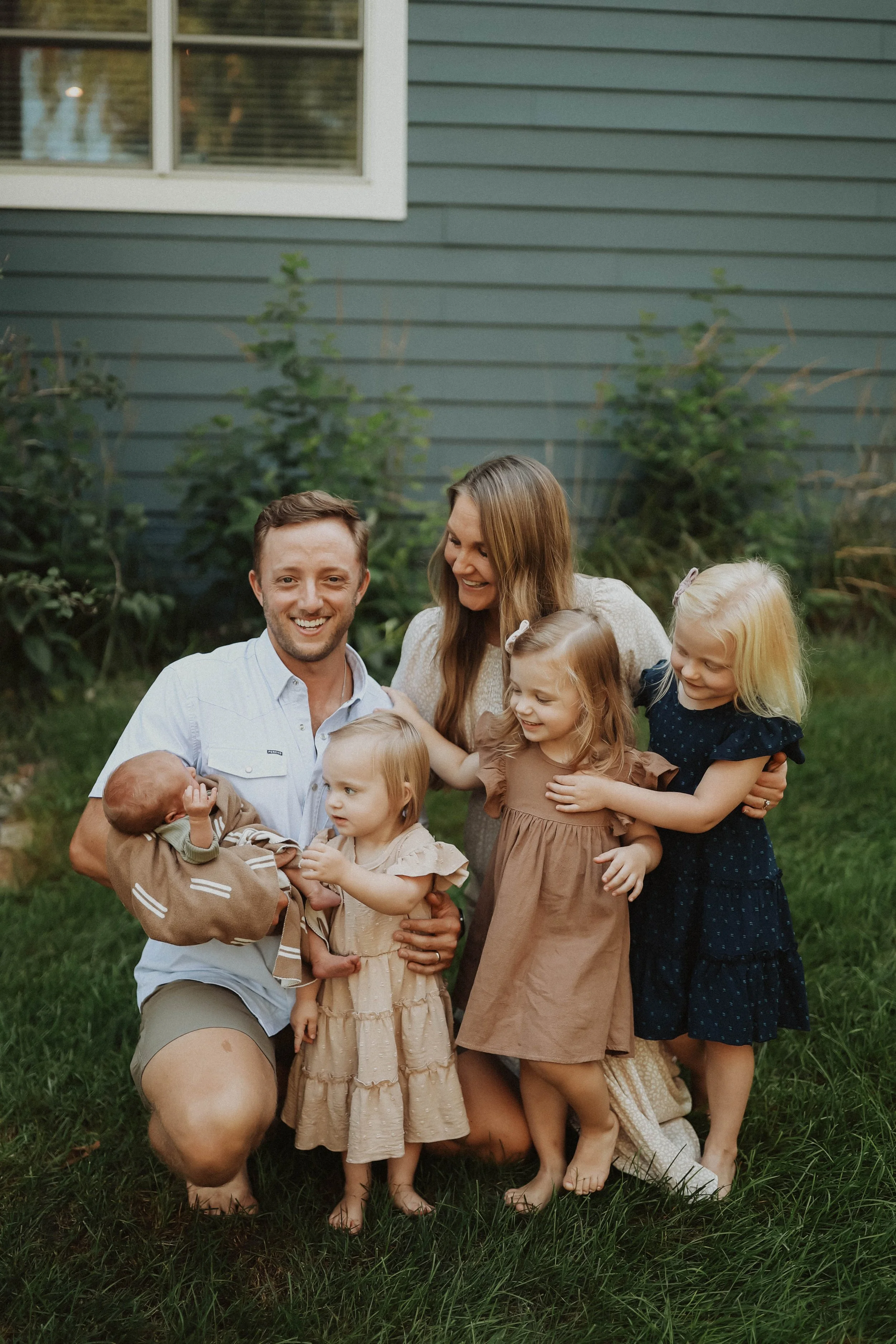 A family of six - welcoming the newest addition to their family, with a house and bushes in the background. The group includes one father, one mother, three young girls, and a baby boy. They are all smiling and hugging each other.