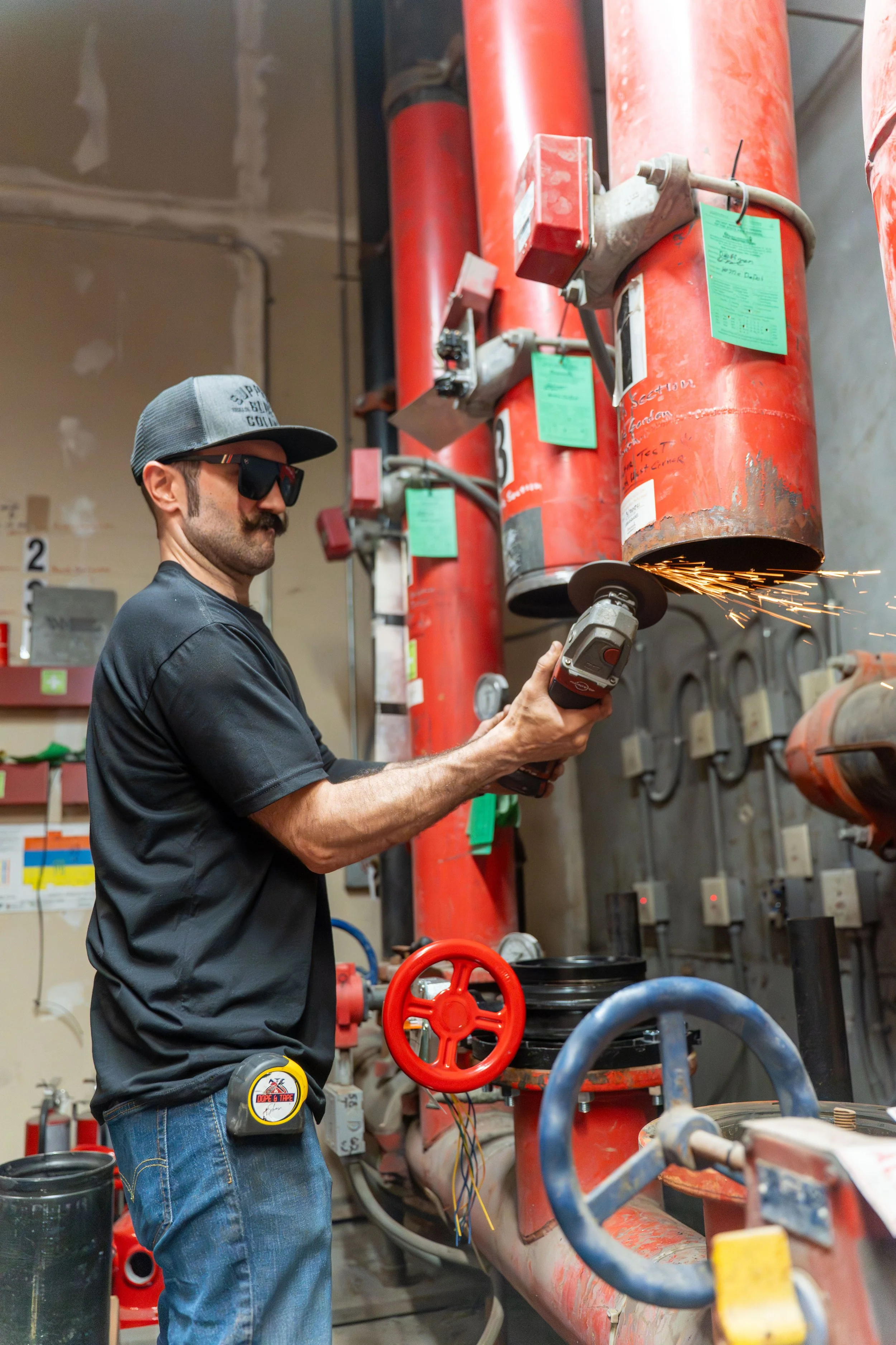 A man in safety glasses and a cap is using a cordless grinder on a large red pipe in an industrial setting, with sparks flying from the pipe's cutting area.