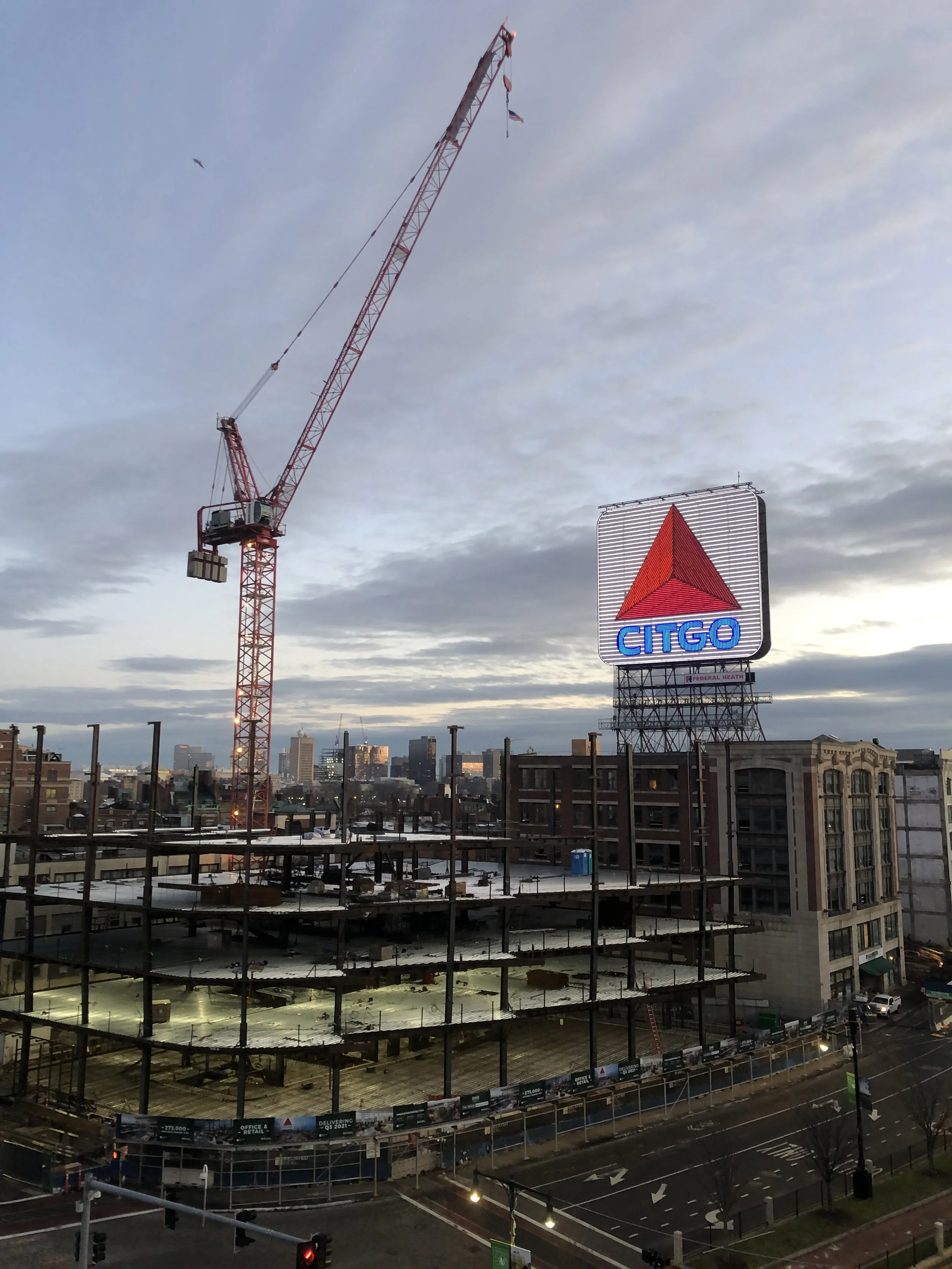 An unfinished multistory building under construction with a large red construction crane and a Citgo sign in the city skyline at dusk.