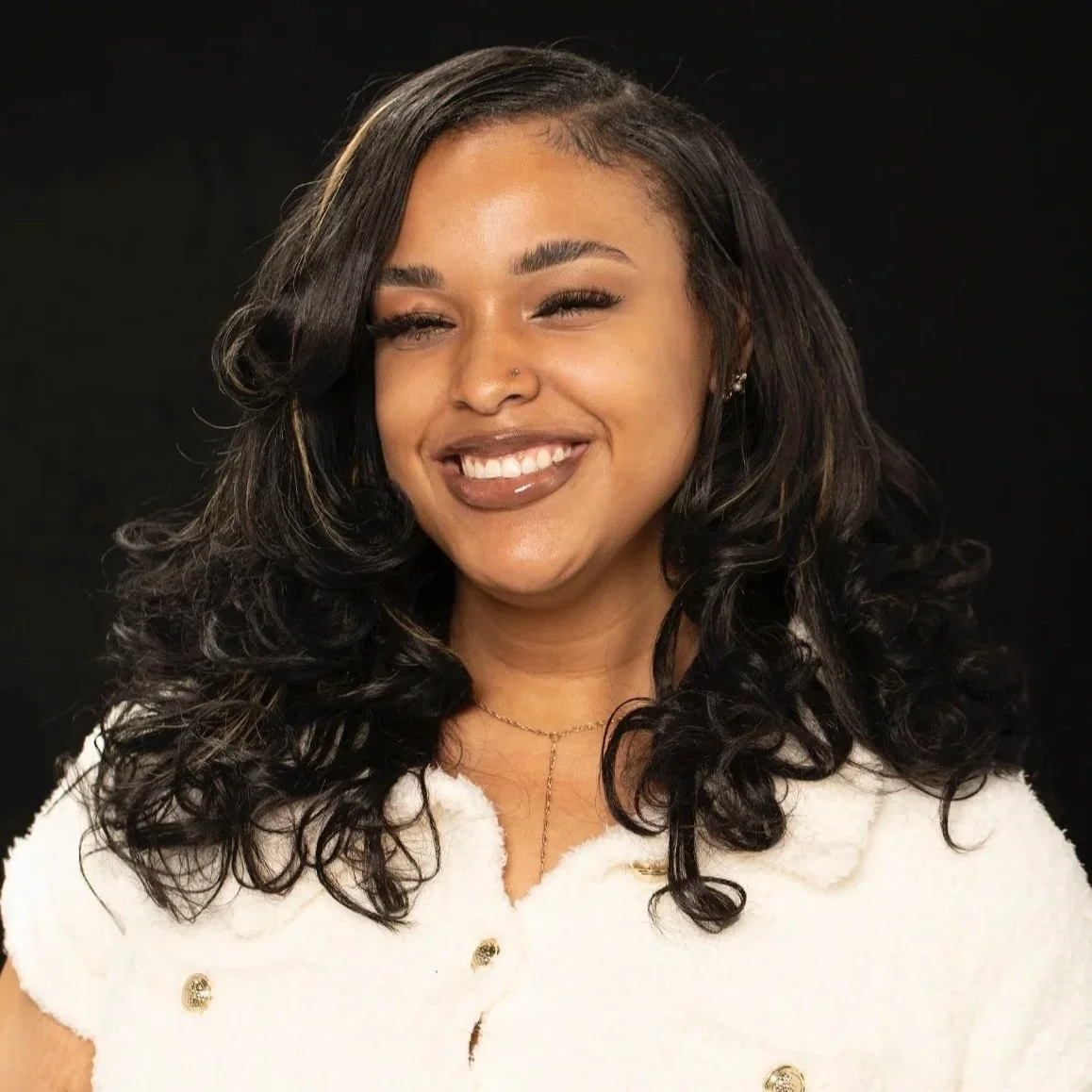 Smiling woman with dark curly hair, wearing a white fur coat and gold jewelry, against a black background.