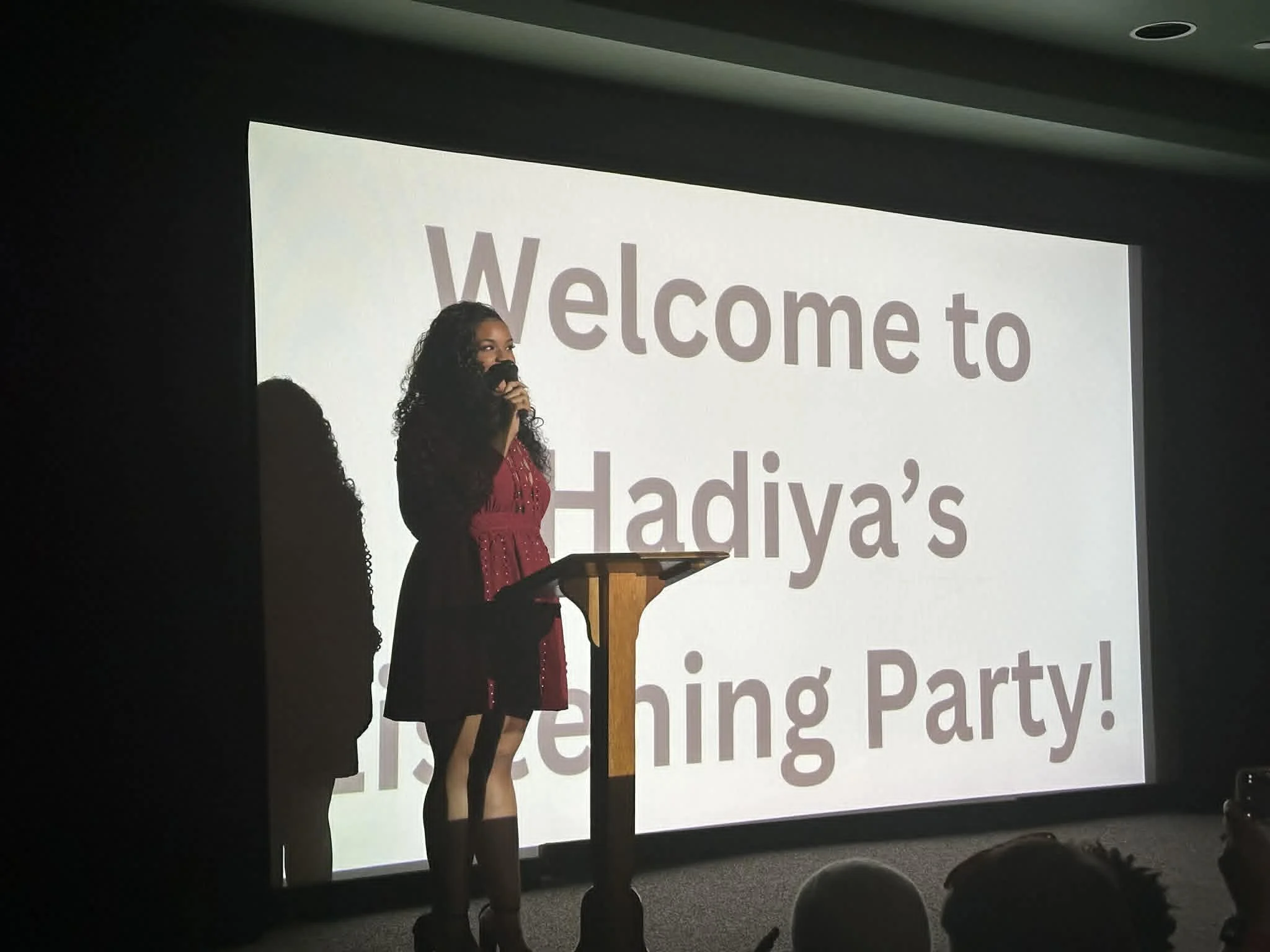 Woman with long curly hair speaking into a microphone at a conference or event, standing behind a podium in front of a large projection screen displaying text.