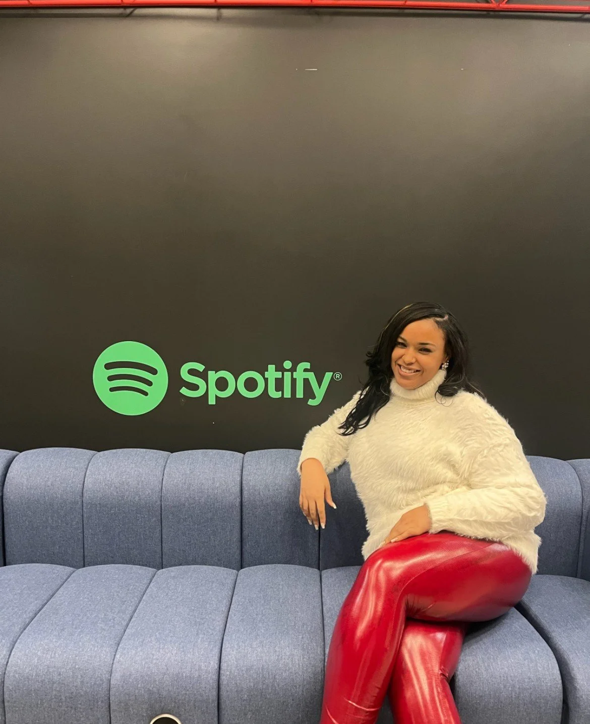 A woman sitting on a blue sofa in front of a black wall with a green Spotify logo. She is smiling, wearing a cream fluffy sweater and red leather pants.