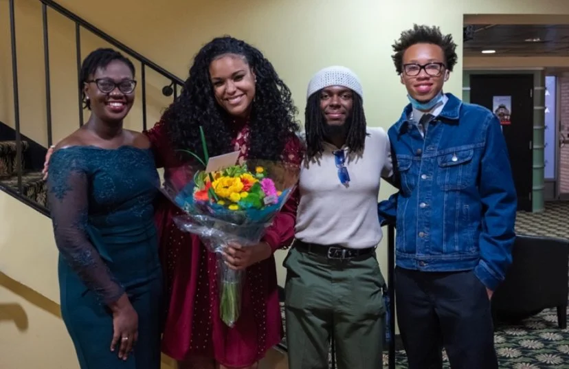Four people standing indoors, smiling at the camera. The second person from the left is holding a bouquet of colorful flowers and a card.