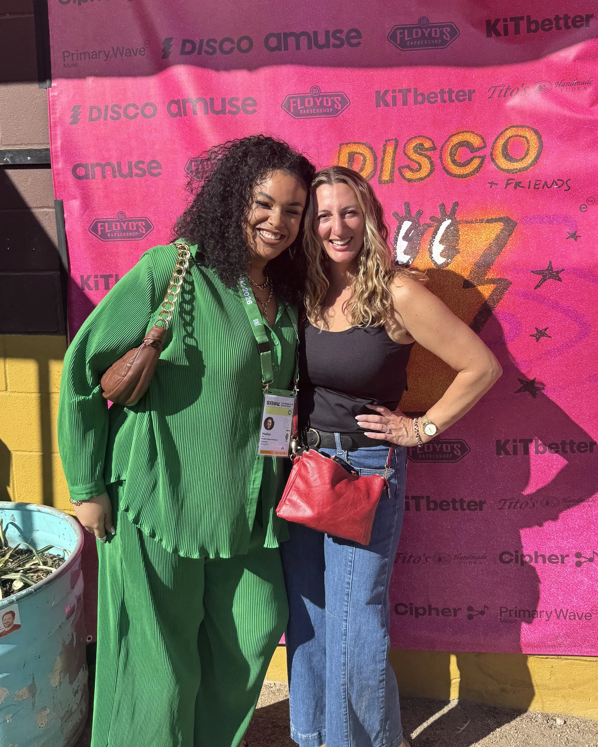 Two women standing close together in front of a pink backdrop with various logos, one woman wearing a bright green dress with a brown purse, and the other wearing a black tank top and blue jeans, both smiling at the camera.