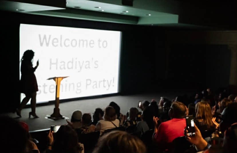 A woman standing on stage with a microphone in her hand, facing a large screen that reads 'Welcome to Hadiya's Testing Party!'. She is silhouetted against the bright screen, with an audience seated in front of her taking photos.