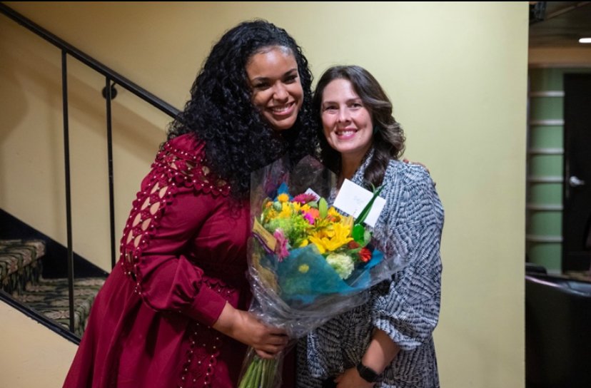 Two women smiling and embracing, one holding a colorful bouquet of flowers, in an indoor setting with a staircase in the background.