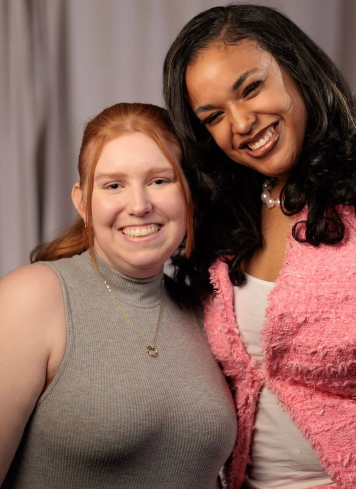 Two women smiling and posing together indoors, one with red hair in a sleeveless top and the other with black hair in a pink jacket.