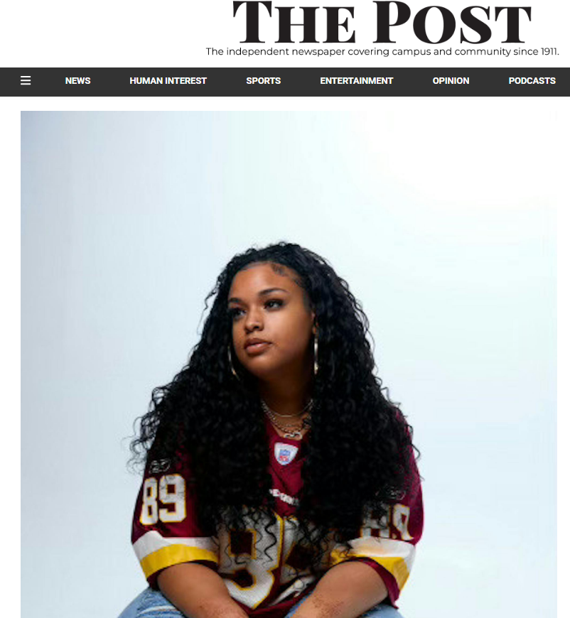 A young woman with long, curly black hair wearing a maroon and yellow football jersey and gold jewelry, sitting against a plain light blue background.