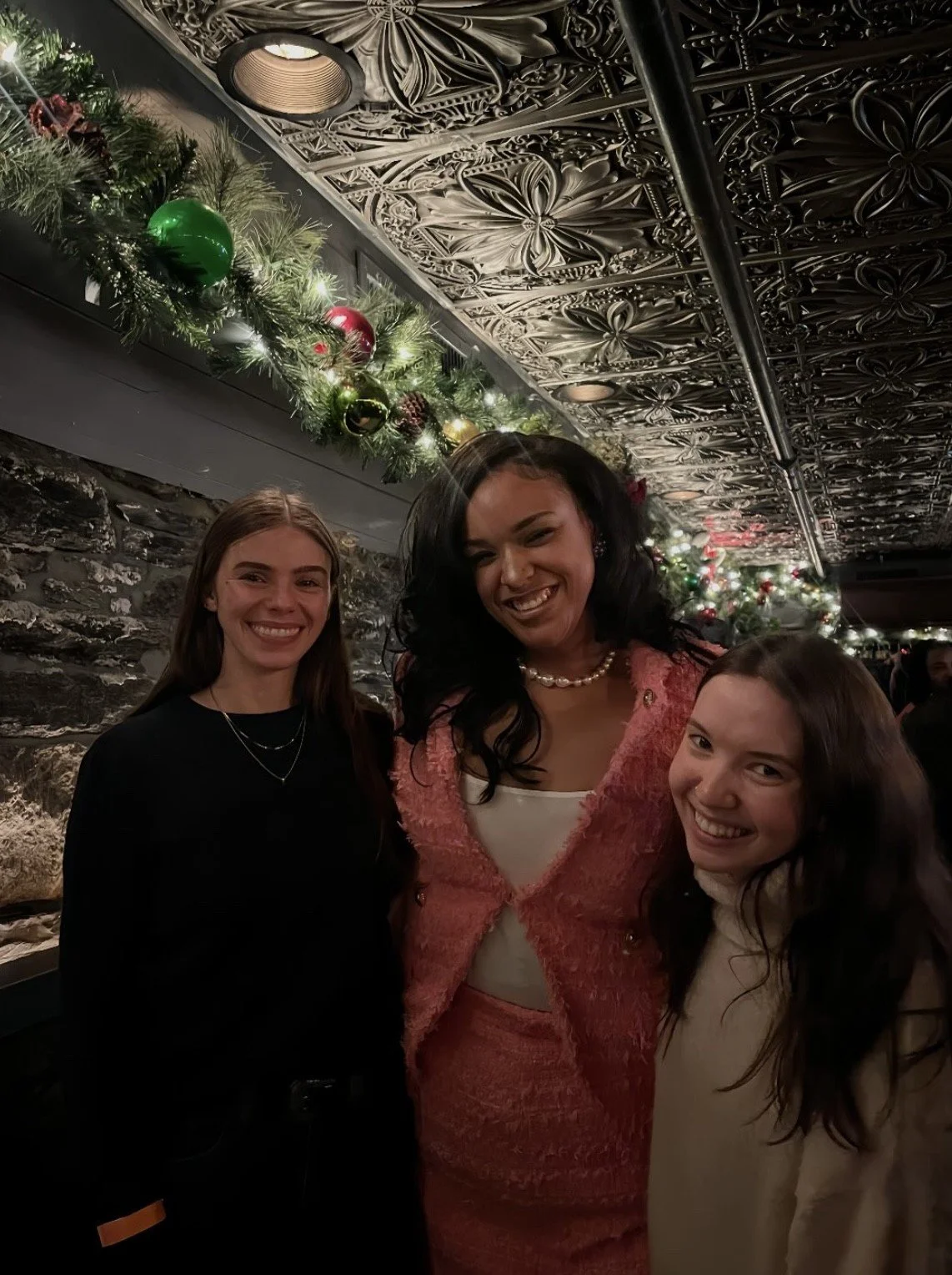 Three women smiling at a Christmas celebration. They are standing in front of a decorated wall with greenery and ornaments, and an ornate ceiling with floral patterns.