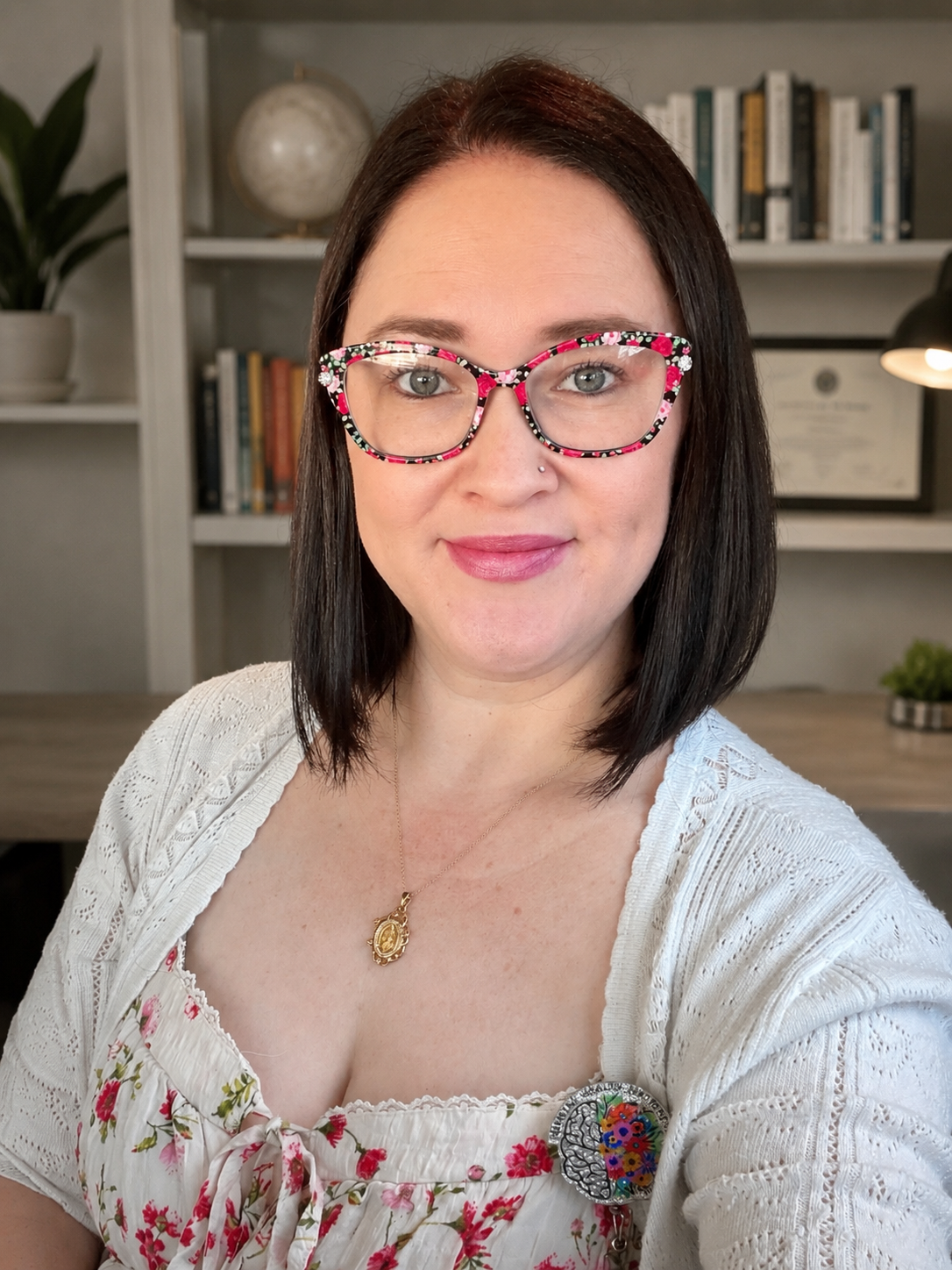 A woman with shoulder-length dark hair and light skin, wearing colorful floral glasses, a white cardigan, and a floral top, taking a selfie in an indoor office or study space with bookshelves and a desk in the background.