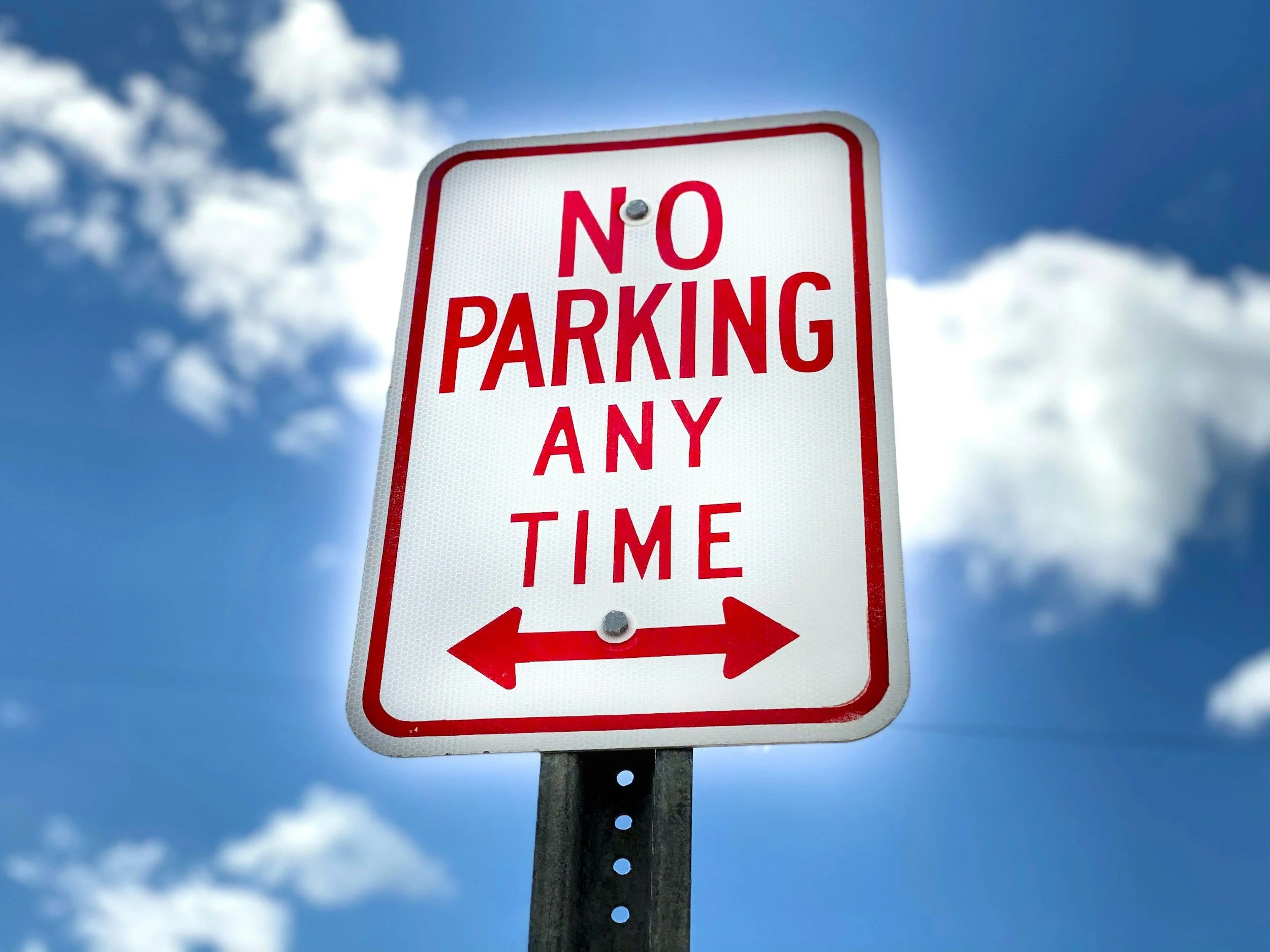 A white and red 'No Parking Any Time' sign with black bolts, mounted on a pole, against a background of blue sky with white clouds.