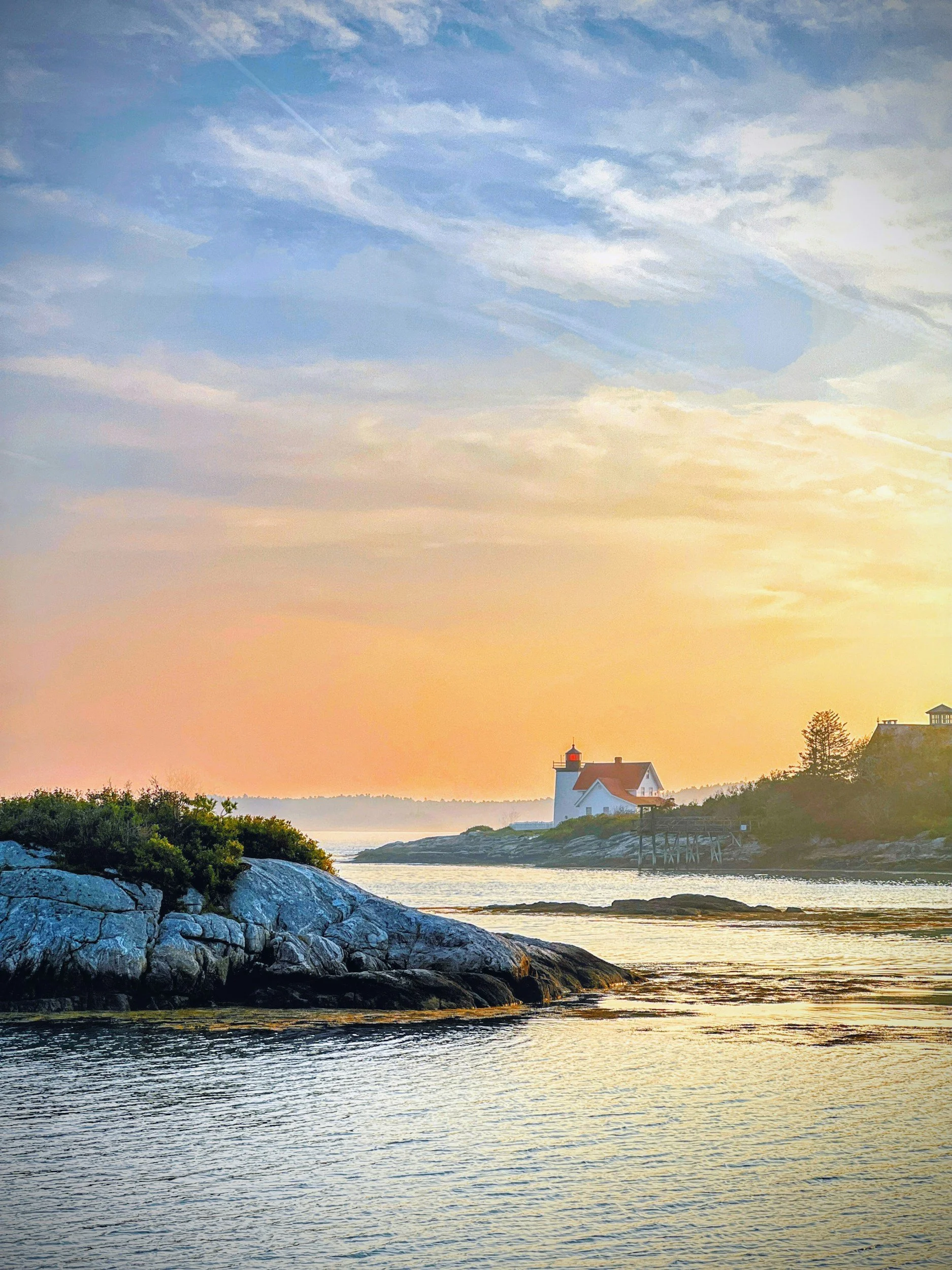 A serene coastal scene at sunset featuring a white lighthouse with a red roof, situated on a rocky shoreline with sparse greenery. The sky displays a gradient of pastel colors, transitioning from pinkish-orange near the horizon to pale blue, with soft, wispy clouds above. The calm water reflects the sky's colors, and there are a few buildings on the distant shoreline.