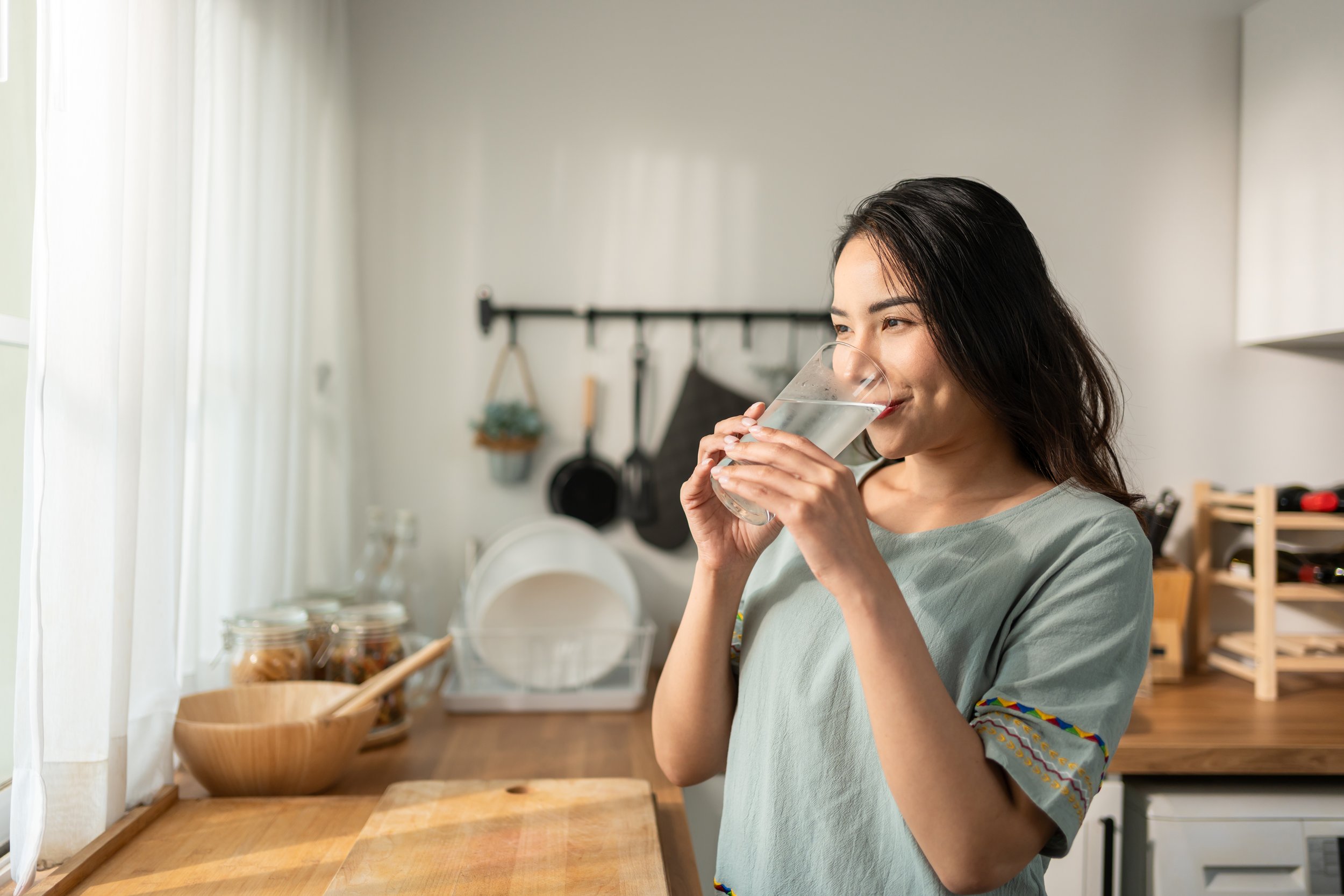 A woman with dark hair drinking water from a glass in a bright, modern kitchen.