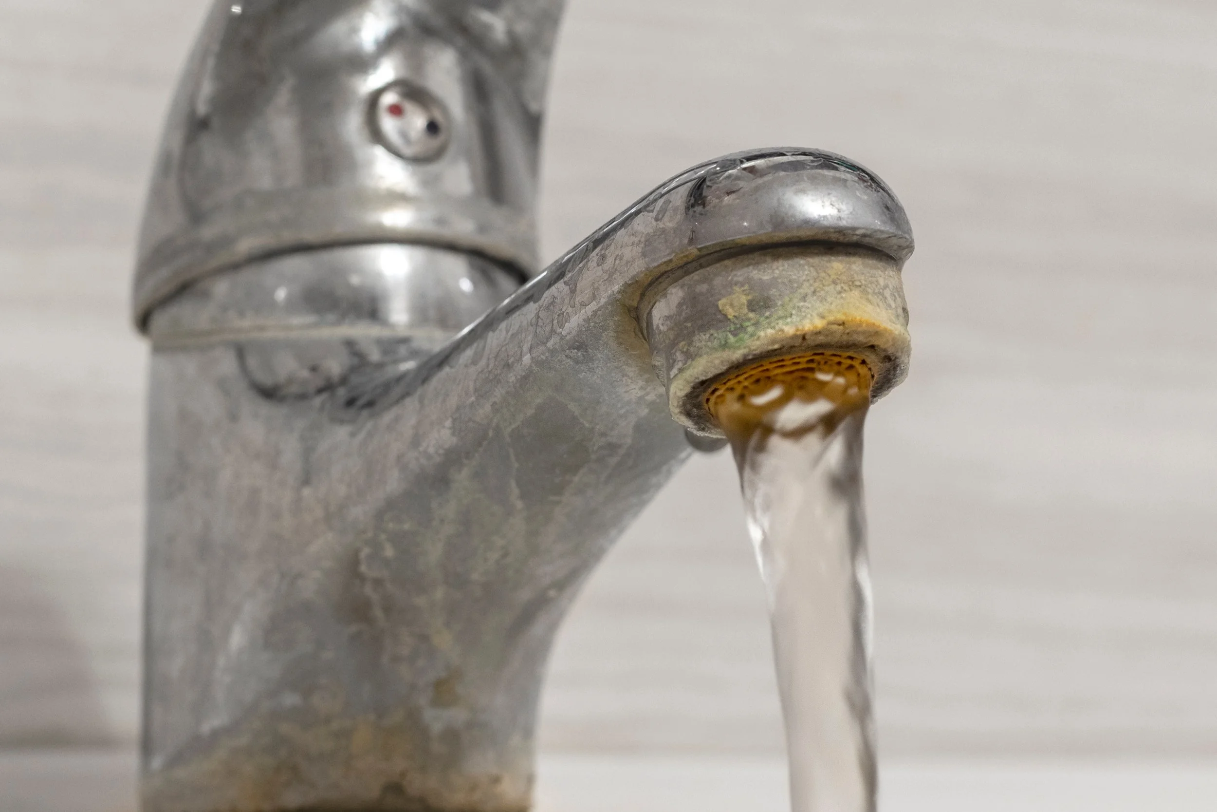 A close-up of a household faucet with water flowing out, showing the metal spout and handle, with a soap or mineral buildup near the base.