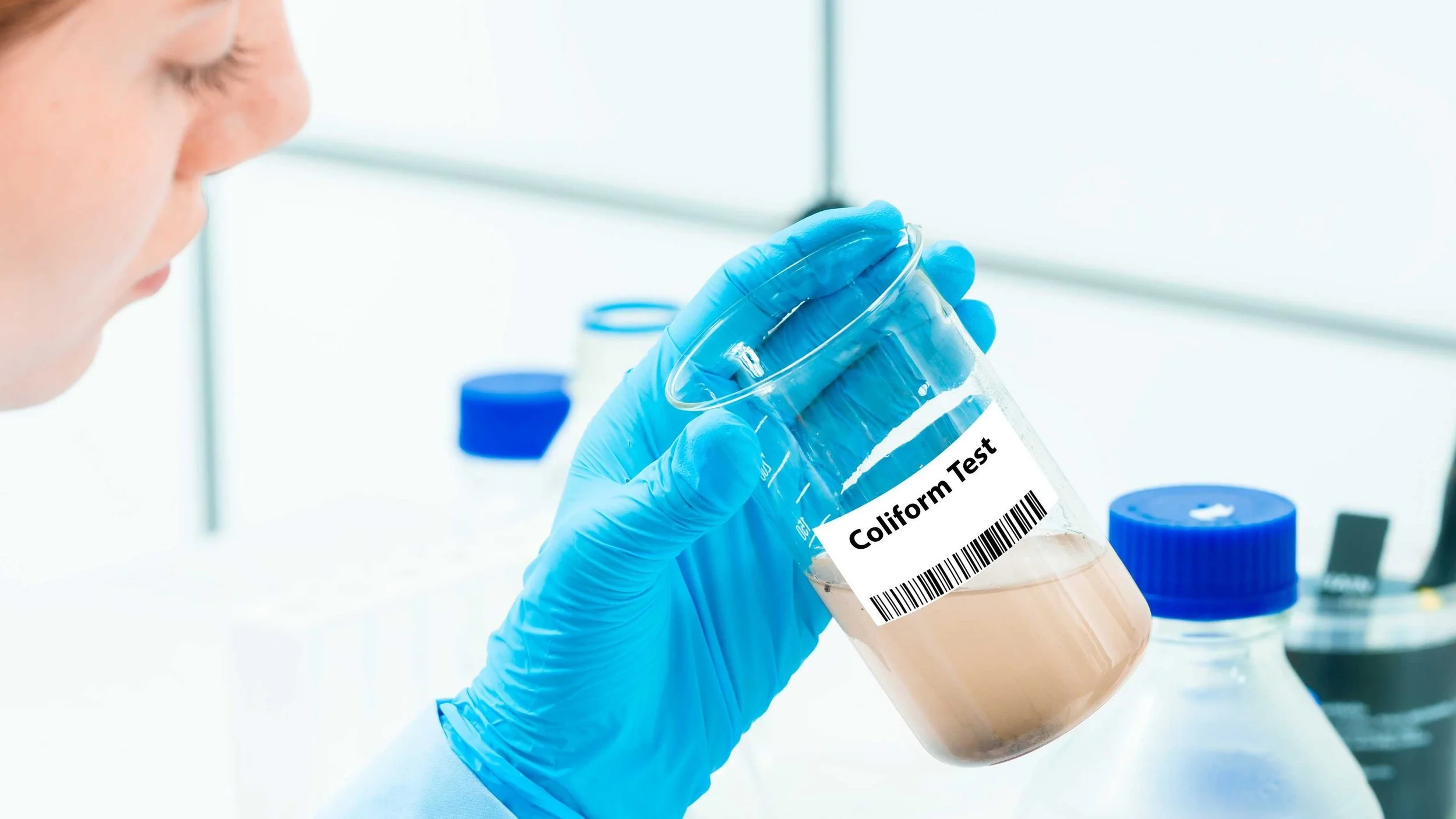 A scientist wearing blue gloves holding a lab test tube labeled 'Coliform Test' with a light-colored liquid inside, in a laboratory setting.