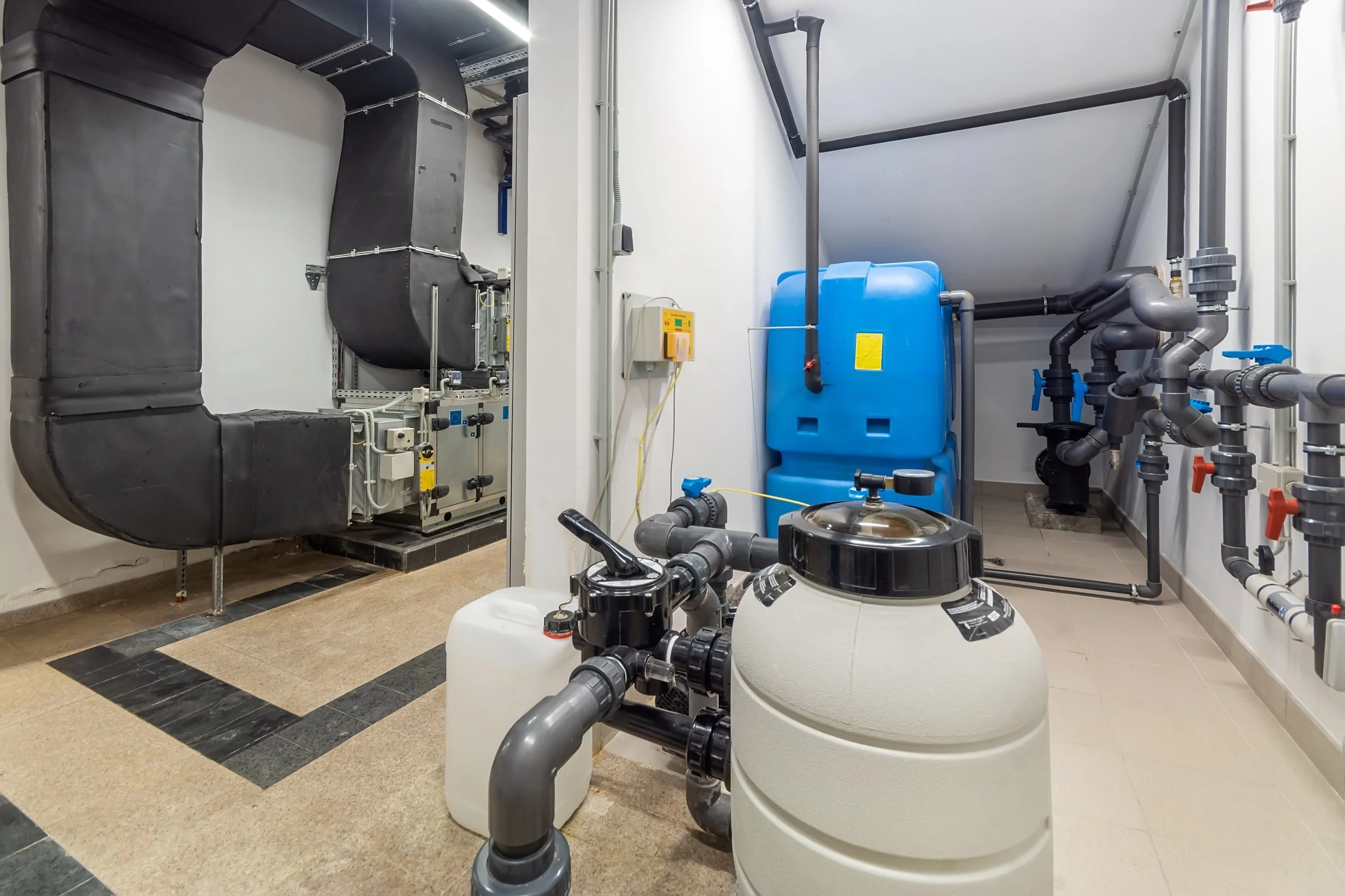 A mechanical room with black ductwork, a blue water tank, and a network of pipes and valves.
