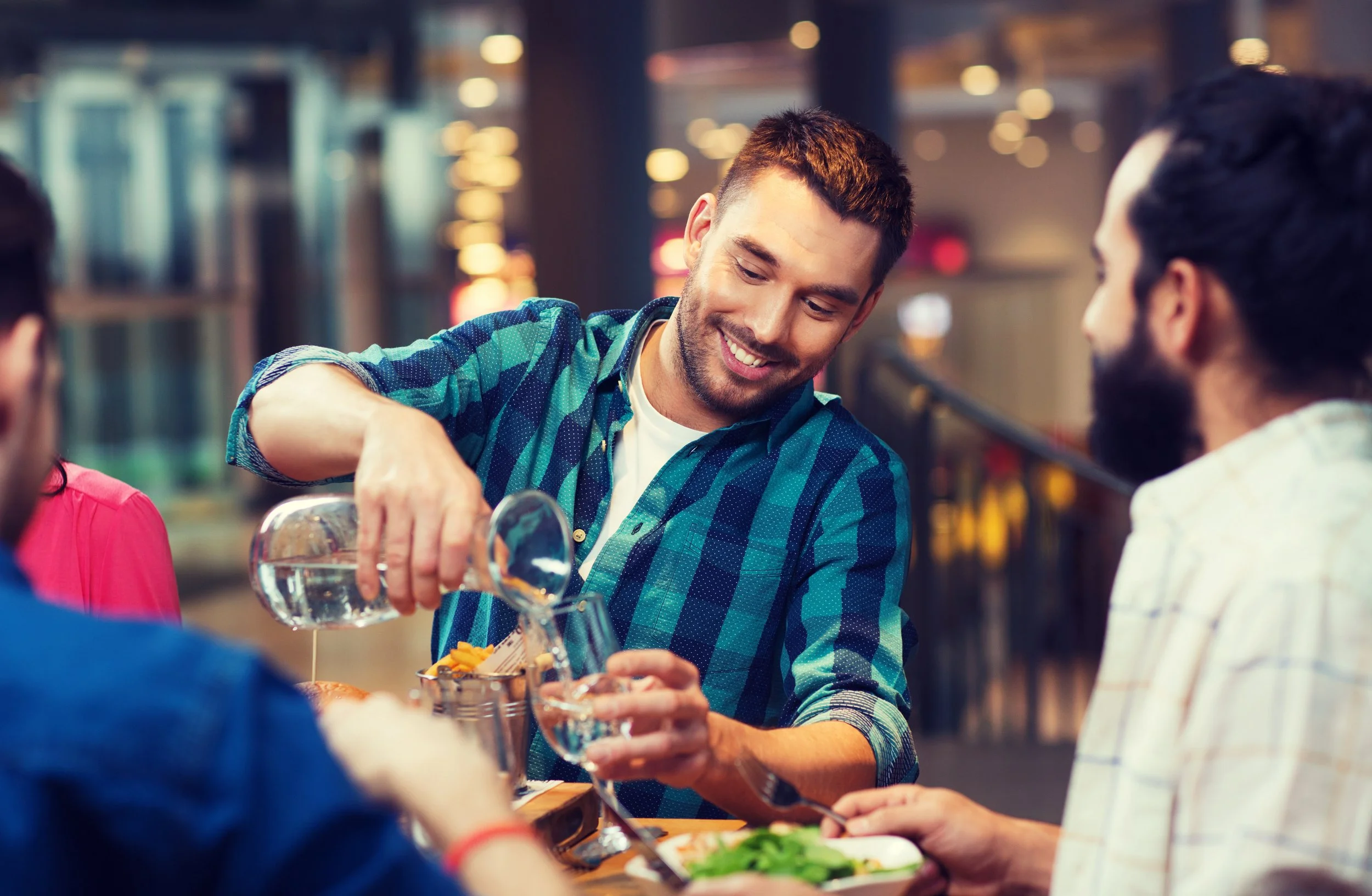 A man in a blue plaid shirt pours water into a glass at a social gathering, surrounded by people, with a blurred background of warm lights.