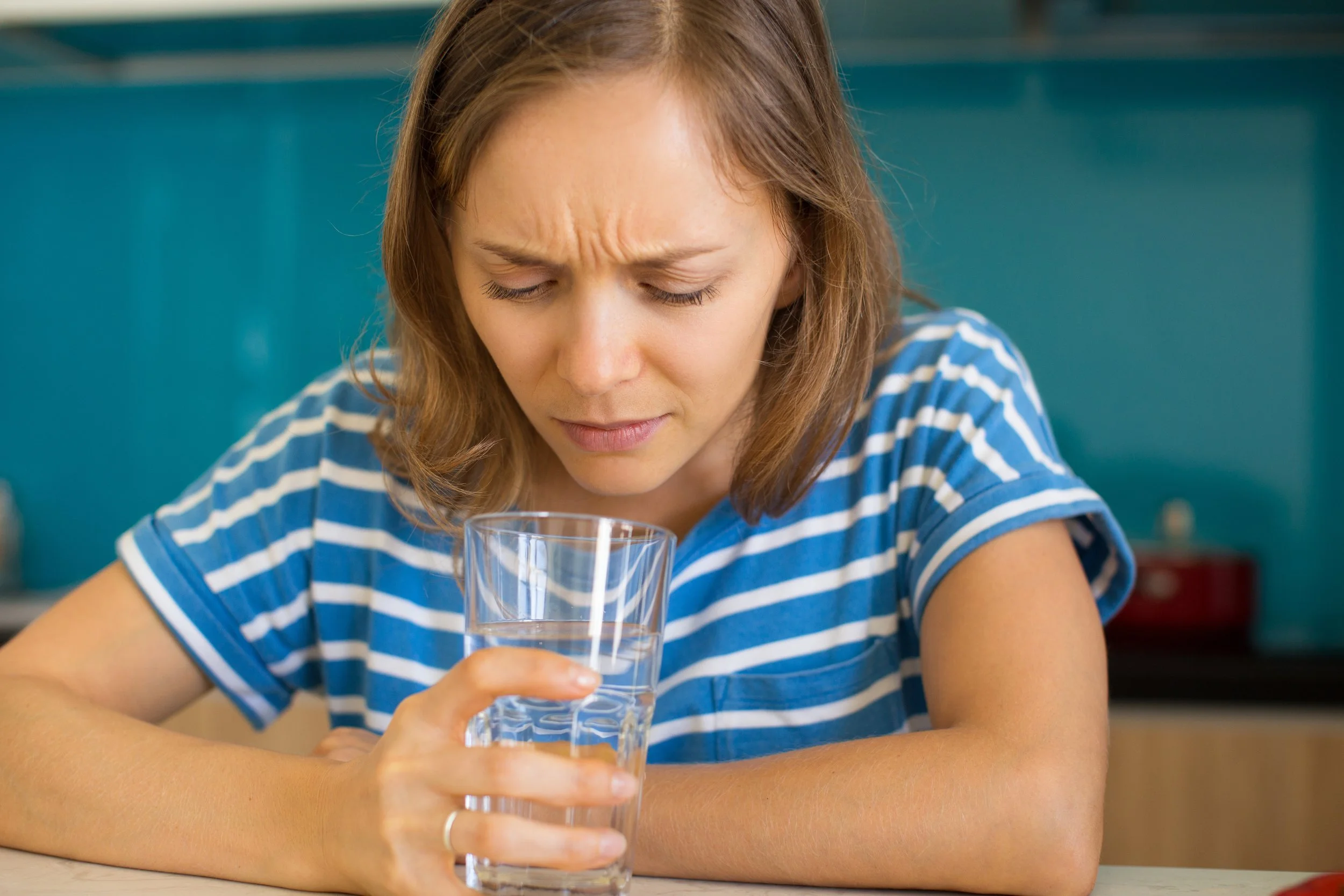 A young woman with brown hair wearing a blue and white striped shirt looks upset or frustrated while holding a glass of water on a table in a restaurant or cafe environment.