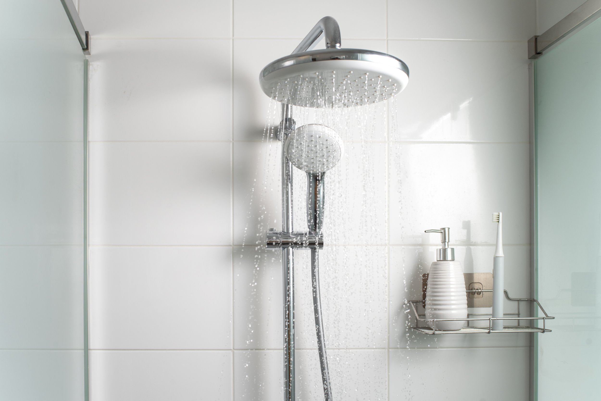 Close-up of a modern showerhead with water running, against a white tiled bathroom wall. On the right, there is a soap dispenser and a toothbrush in a holder.