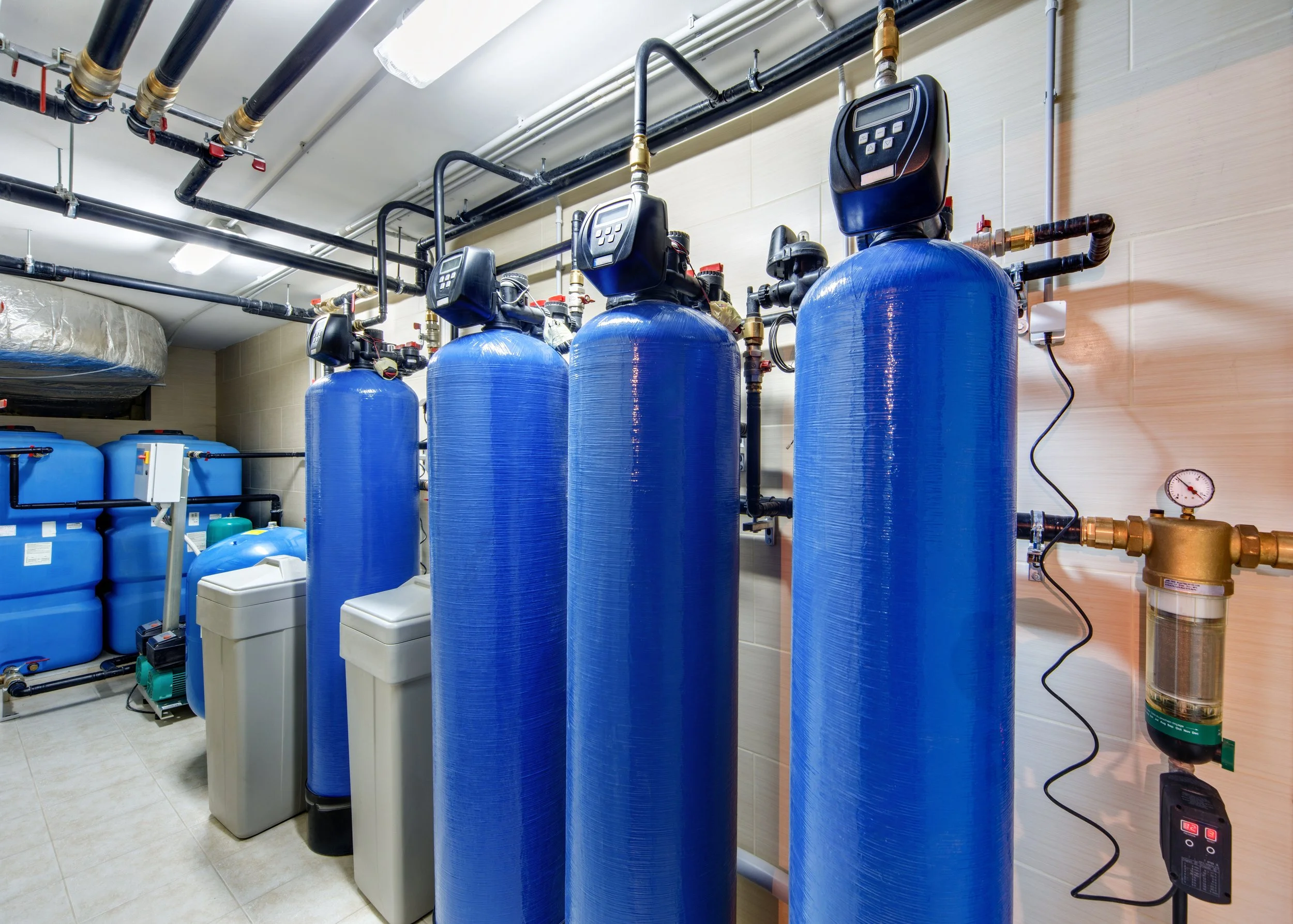 Room containing large blue water filtration tanks, black pipes on ceiling, and control panels on tanks.