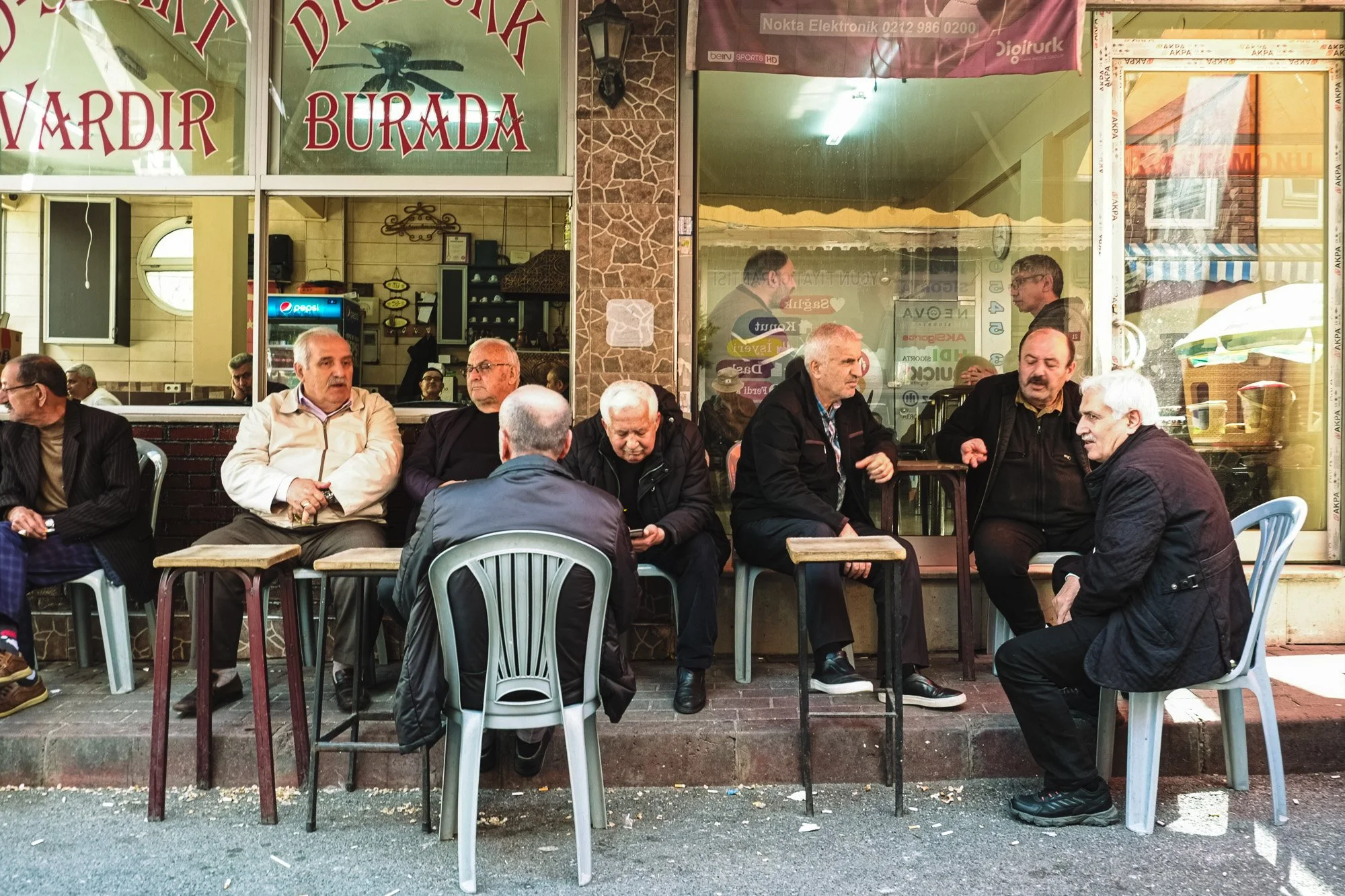 A group of older men sitting outdoors at a cafe or restaurant with plastic chairs and tables, engaged in conversation and looking in various directions. The establishment behind them has large glass windows, showing the interior with more people and 
