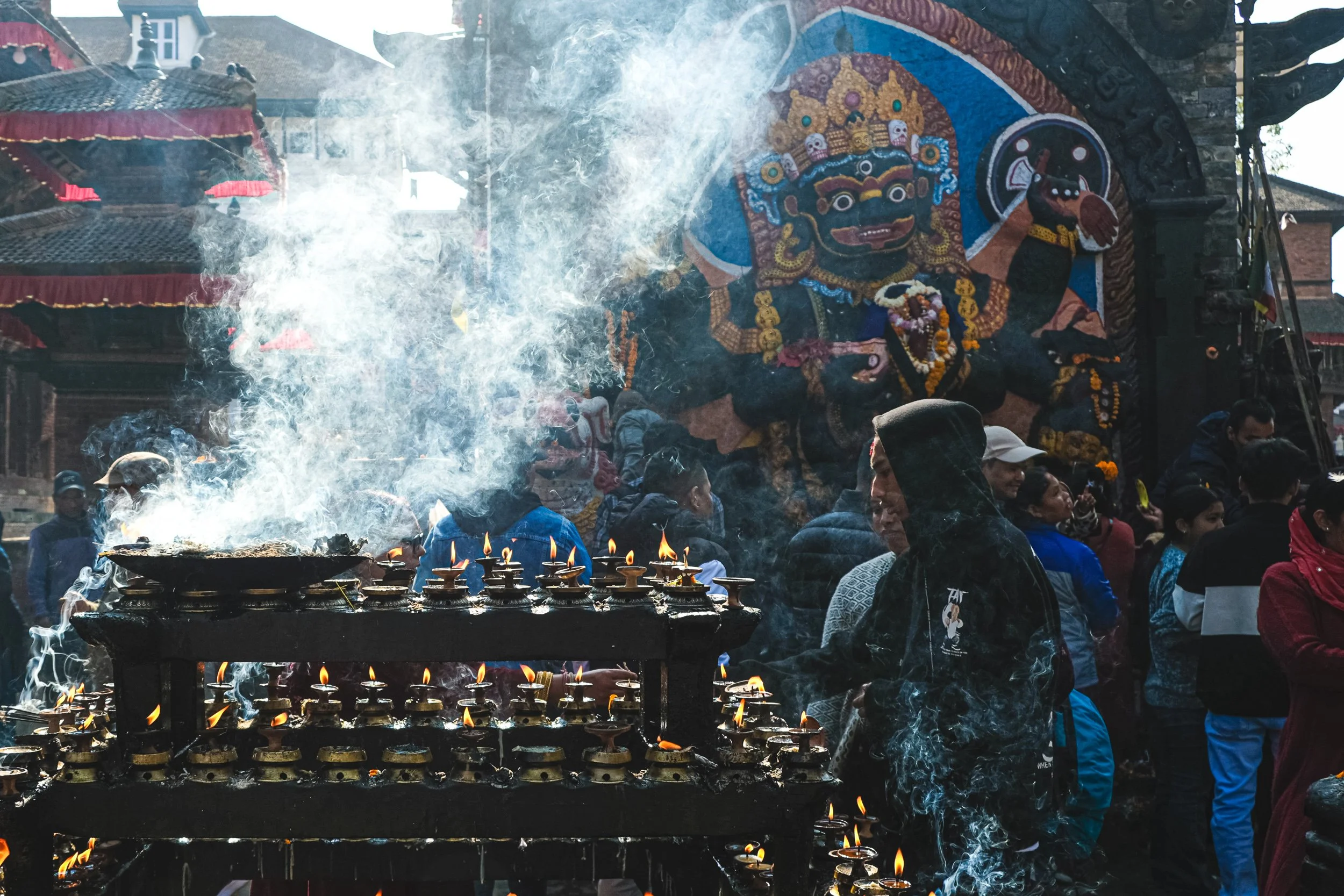 People gathered in a crowded outdoor space during a cultural or religious celebration. The scene includes a large, colorful statue of a deity or mythological figure, decorated with intricate details and garlands. Several lit oil lamps are placed on a