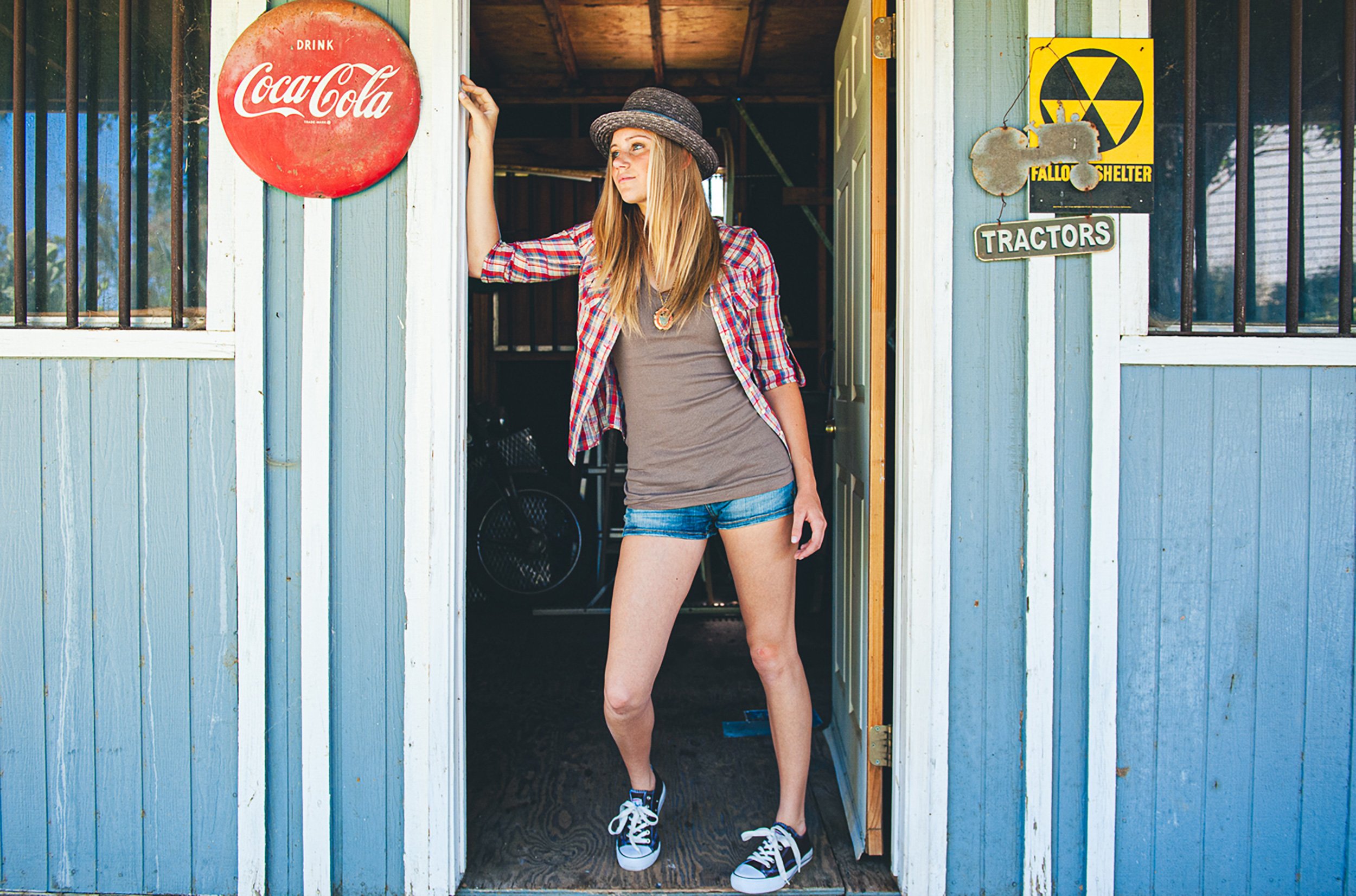 A young woman is standing at a door of a blue wooden shed, wearing a plaid shirt, gray t-shirt, denim shorts, sneakers, and a straw hat. She is looking to her left, with her right arm raised and holding the door frame. The shed has barred windows and