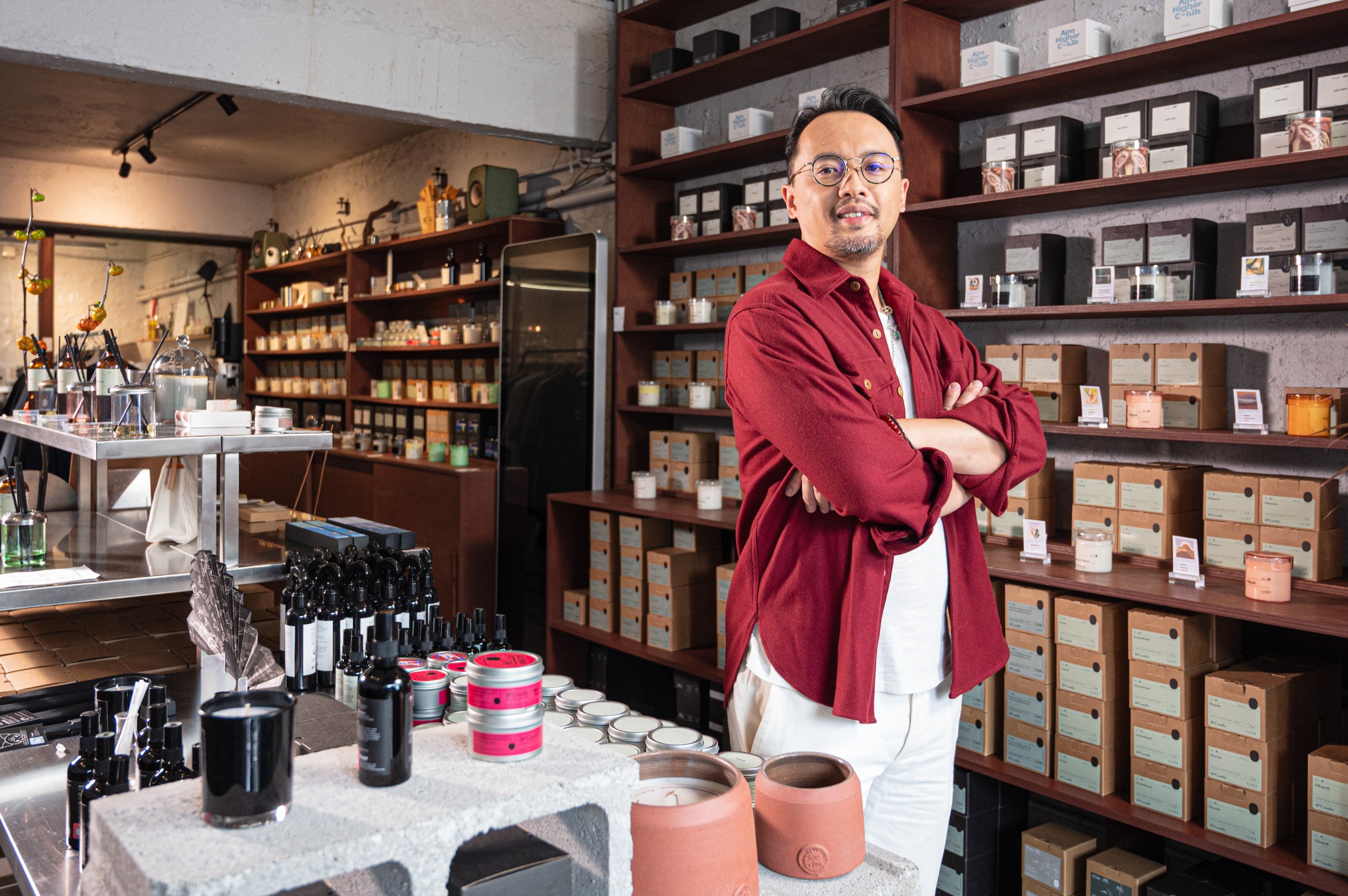 A man wearing glasses, a red jacket, and white pants standing with arms crossed inside a store with shelves filled with boxes and jars, and a table displaying bottles and containers.