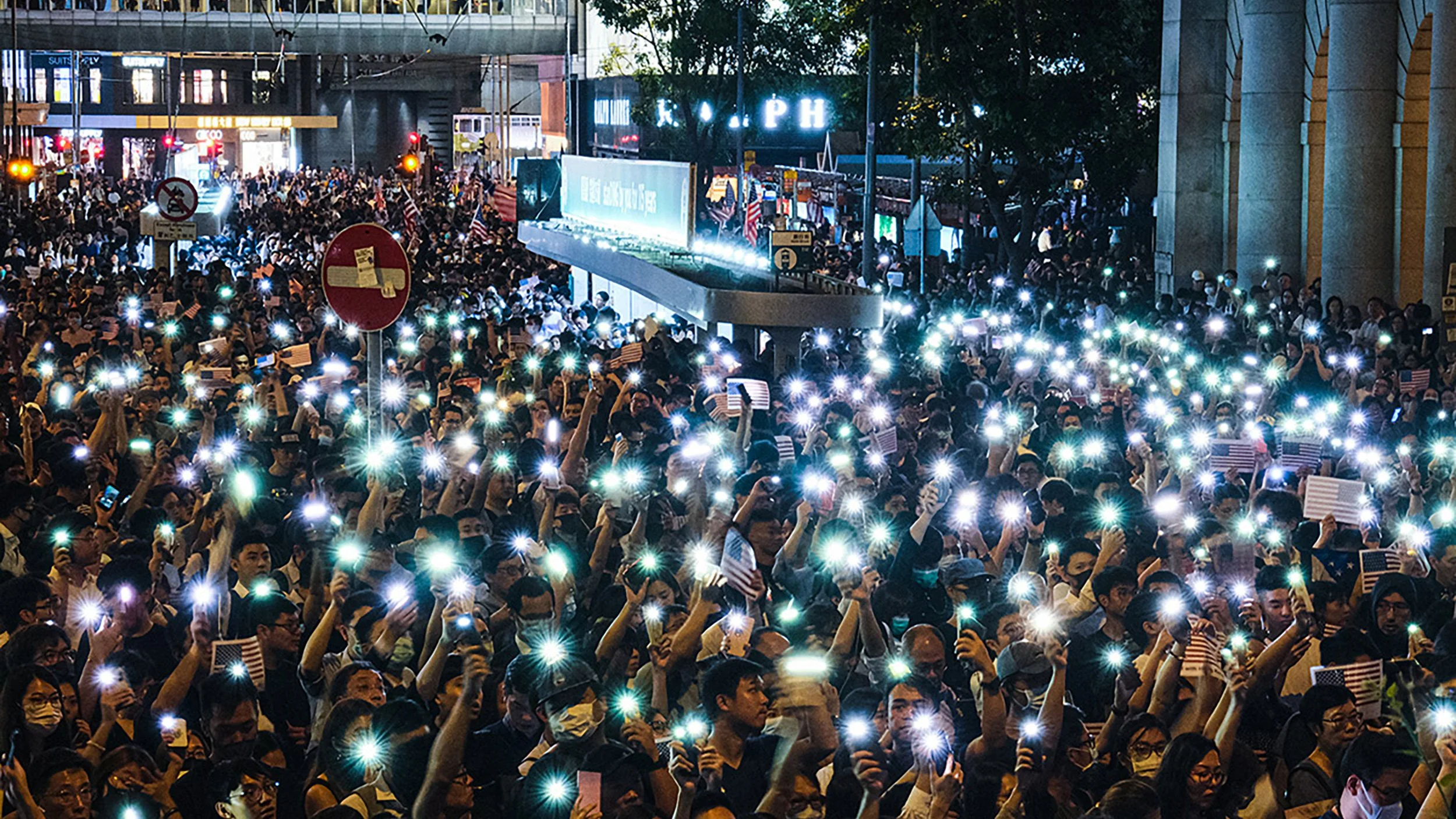 Crowd of people at a nighttime protest or rally holding up illuminated phones and American flags, with signs and buildings in the background.