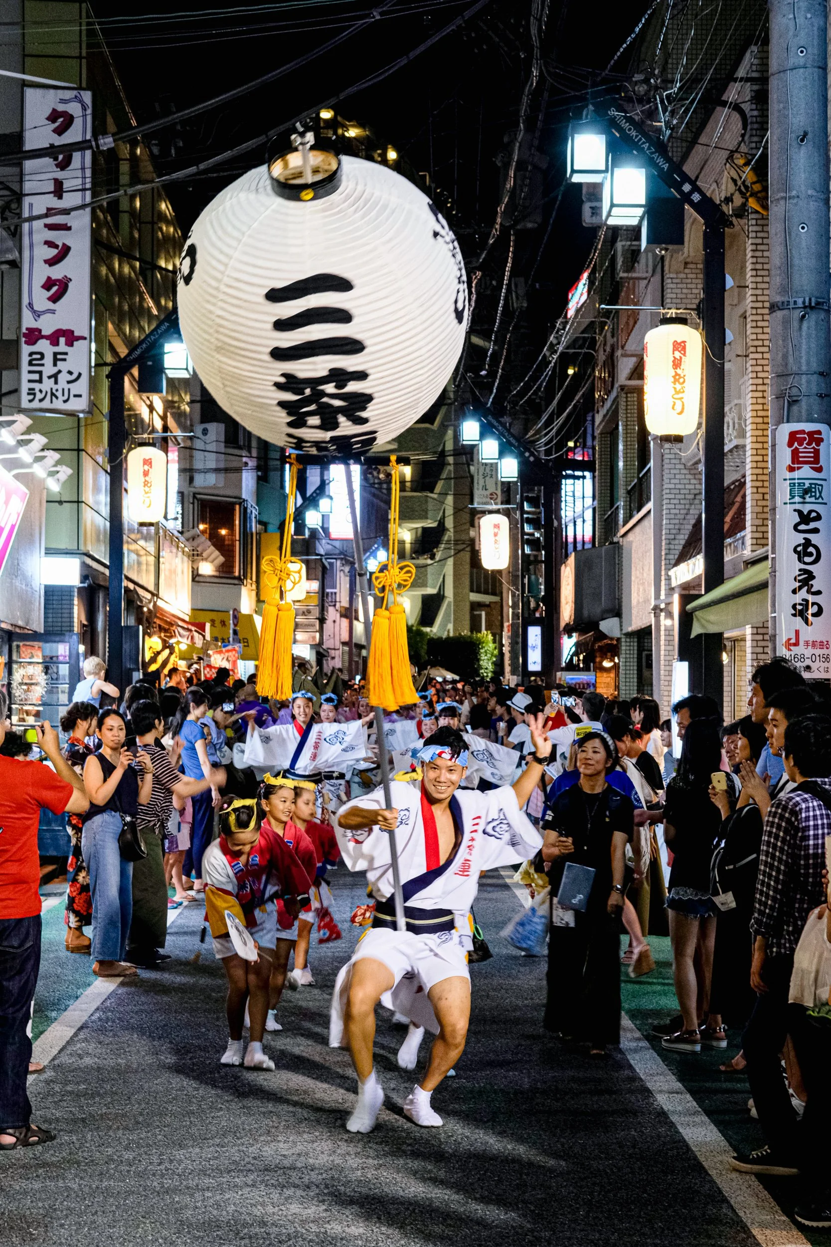 Street scene at night during a festival with a procession of performers in traditional Japanese clothing, including children and adults, with lanterns hanging overhead and a large lantern with Japanese characters in the foreground. Crowds of spectato