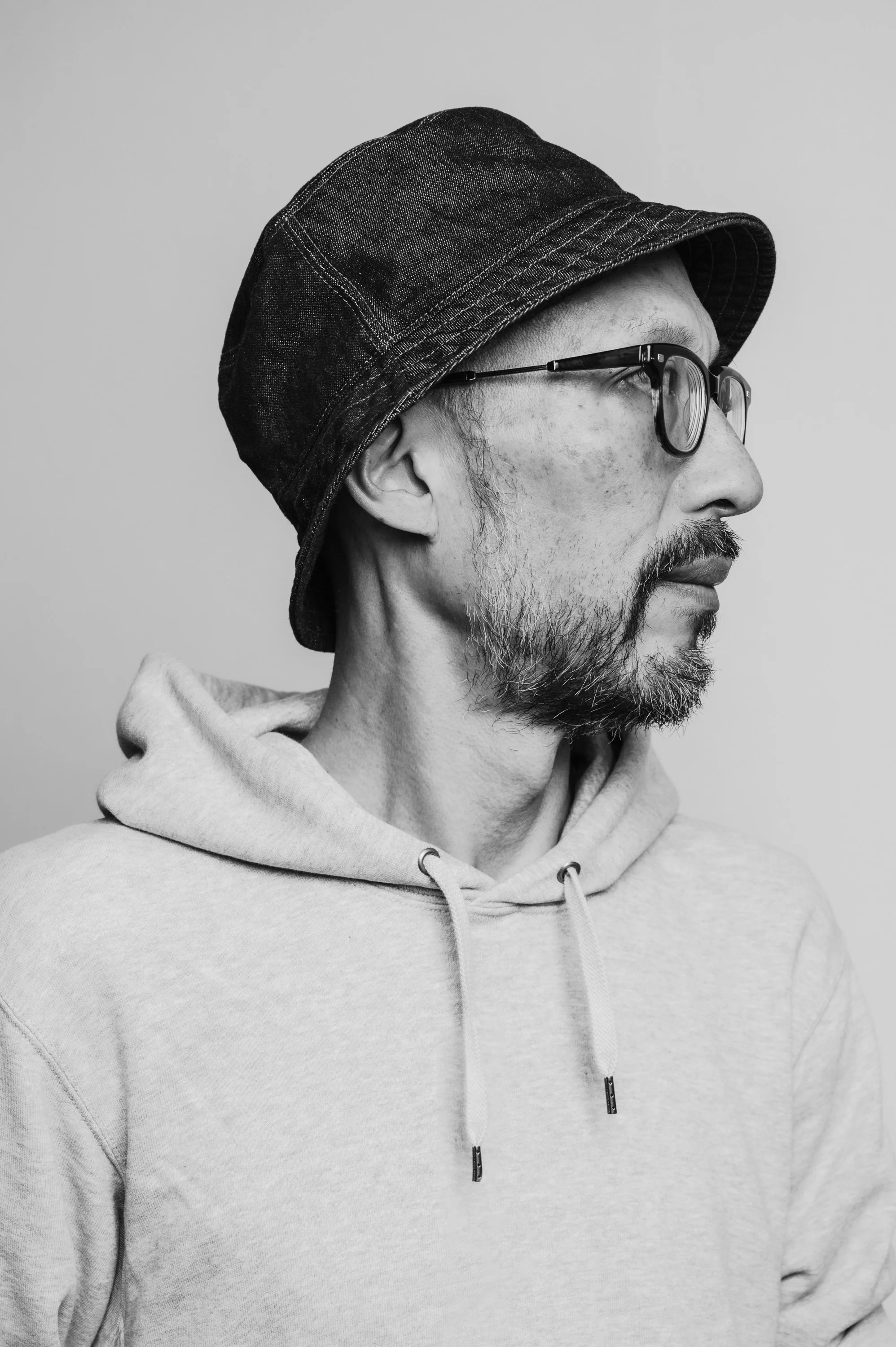 Side profile of a man wearing a dark denim bucket hat, glasses, a beard, and a light-colored hoodie.