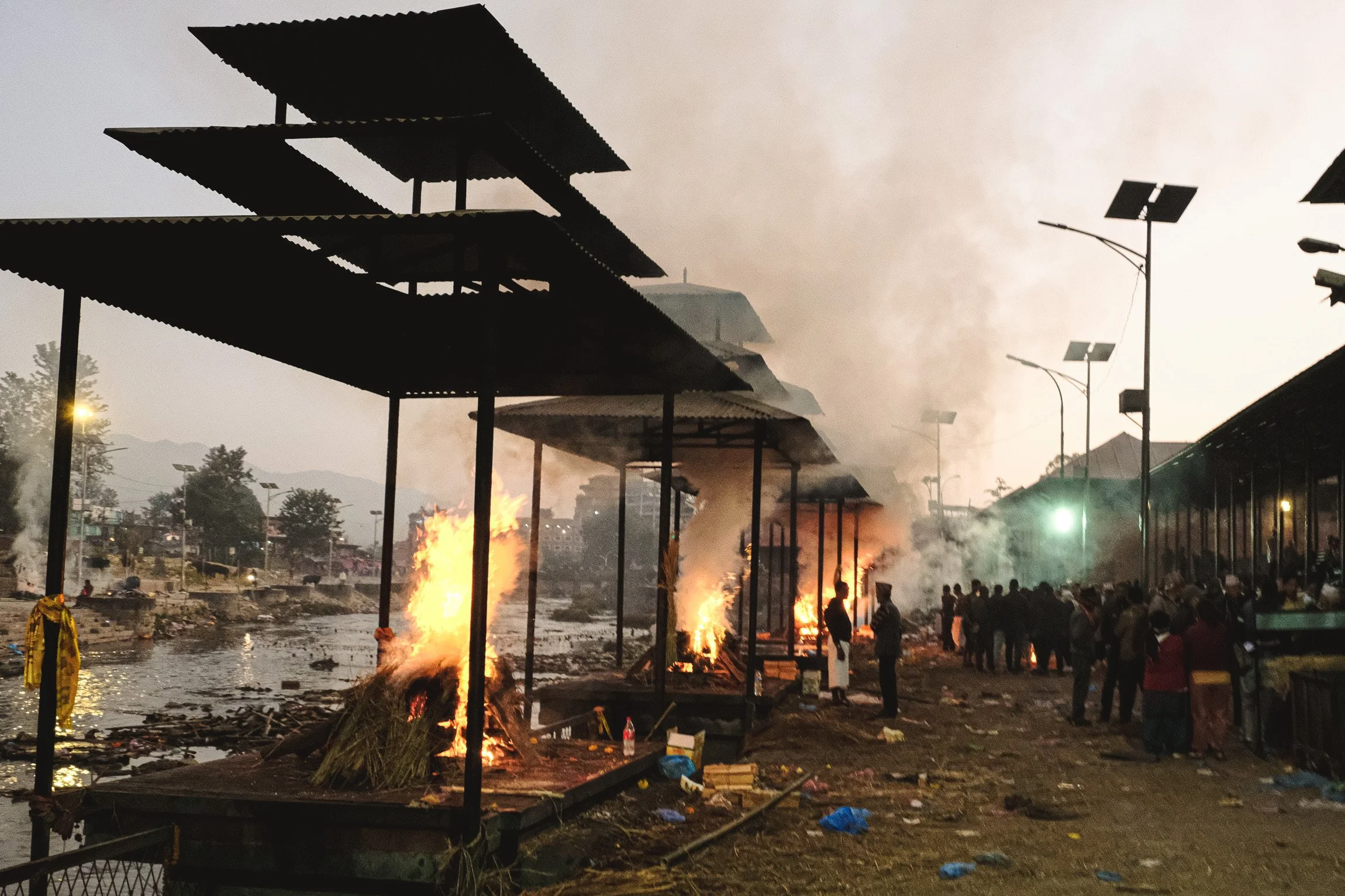 A scene of a fire at a marketplace or outdoor area with smoke in the sky. Several stalls or structures are on fire, and a group of people are gathered nearby, with some observing the incident.
